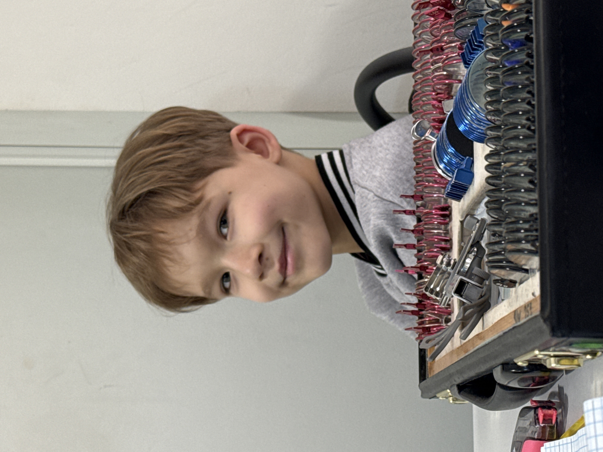 A young boy with short brown hair and a grey sweater with black and white stripes on the collar is looking up and smiling slightly. He is positioned next to a complex object with many small springs of red, silver, and blue metal. The object also contains small metallic tools. The background is a plain light grey wall. There is no information about the location in Kuleshovka, Russia.