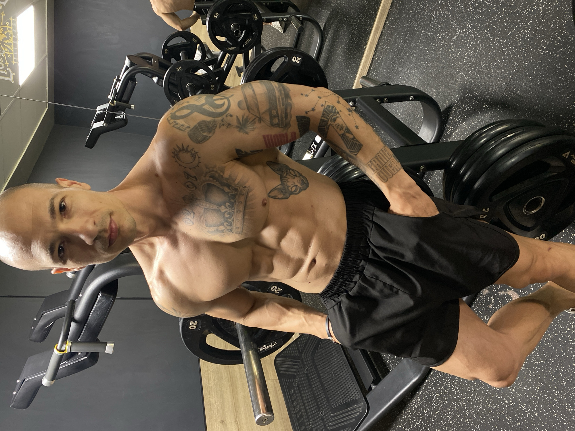 A man with visible tattoos on his arms and chest poses in a gym setting. He is wearing black shorts and is shirtless, displaying his physique. Gym equipment, including weight plates, is visible in the background. The photo appears to be taken indoors with a mix of grey and wooden surfaces. There is no indication of the location being Pleven, Bulgaria.