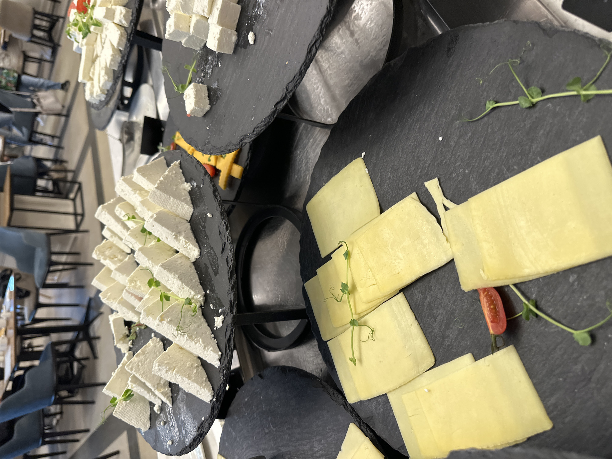The image shows a food display, likely a buffet or catered event. Several multi-tiered black slate serving stands hold a variety of cheeses. One stand displays cubed white cheese, another features triangle-cut white cheese, and a third has sliced yellow cheese, garnished with small green sprouts. A few tomato slices are interspersed with the cheese. The background reveals a restaurant or dining area, with chairs and tables visible. It is unknown if the location is in the city of Kazanlak, Bulgaria, based on this image.
