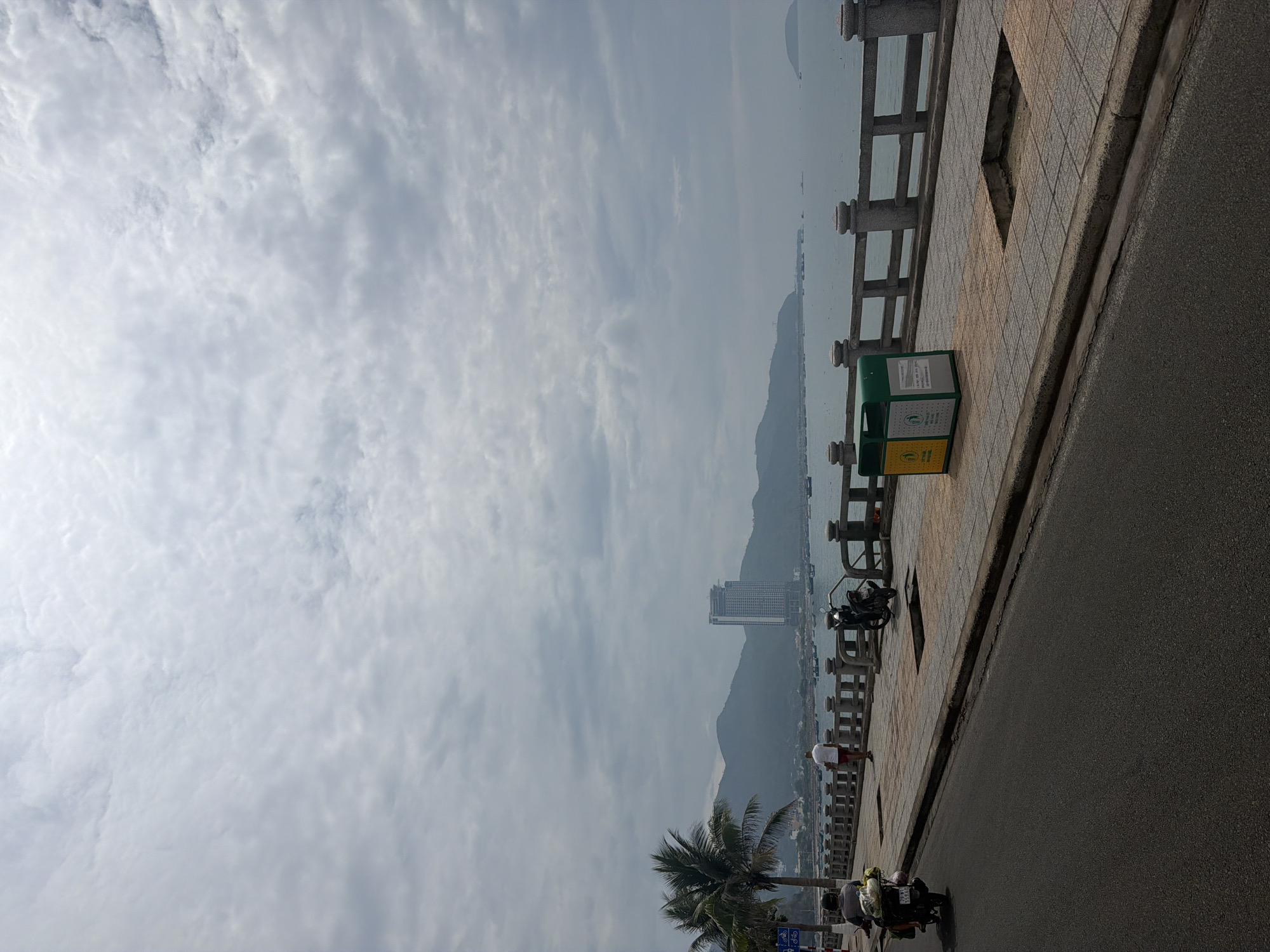 The image shows a view in Bac Nha Trang Ward, Vietnam. The sky is overcast with numerous clouds. In the background, mountains are visible along the coast, obscured by haze. A tall building is present amidst the landscape.
The foreground consists of a sidewalk with a concrete balustrade overlooking the water. Adjacent to the sidewalk is a street with dark asphalt. There is a trash bin on the sidewalk.
A person is walking along the sidewalk. A motorcycle with two riders is on the road. Parked motorcycles are seen near the balustrade. A palm tree is also visible.