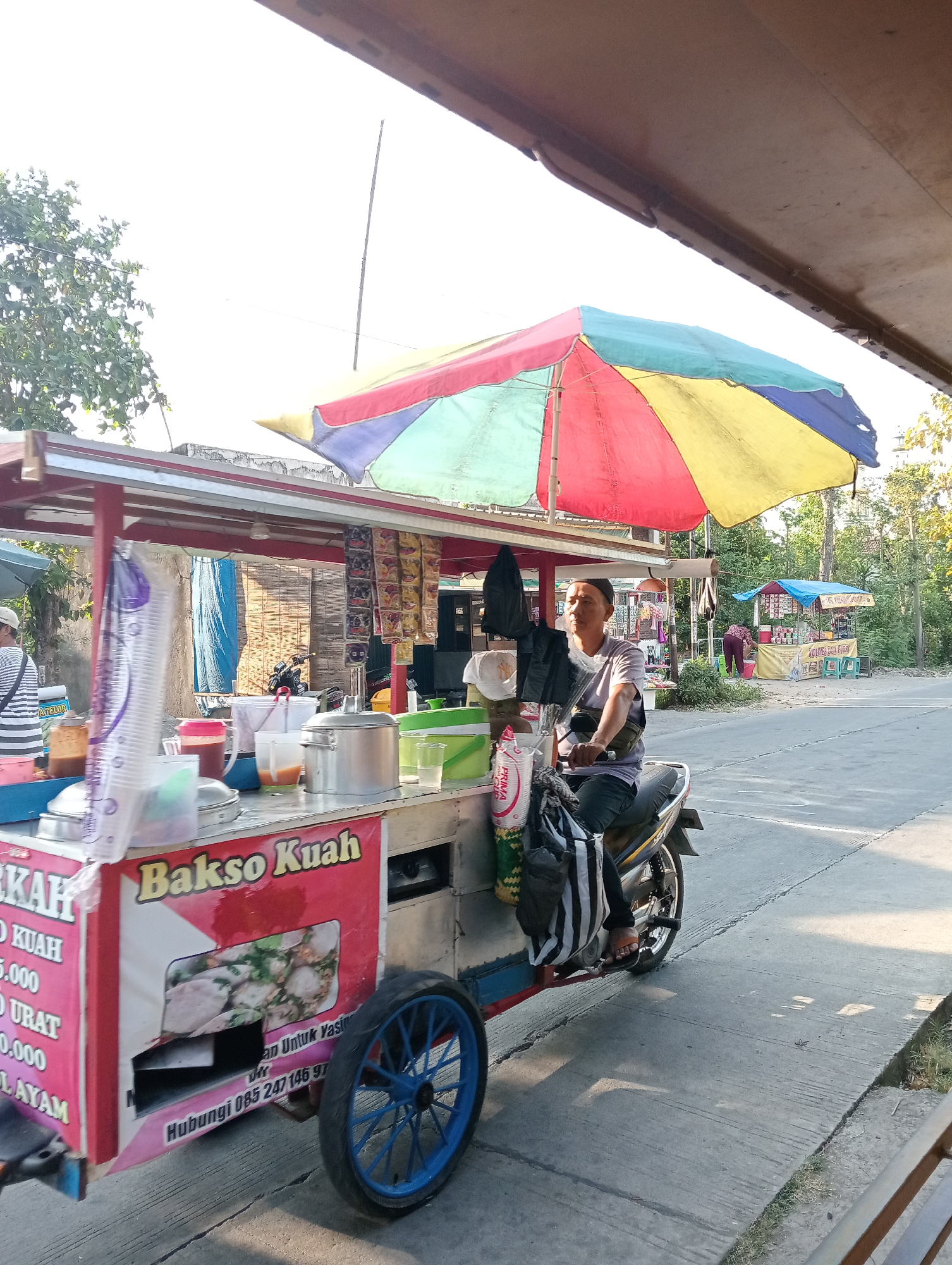 In Tanjunganyar, Indonesia, a man sits on a motorcycle attached to a food cart. The cart, adorned with a colorful umbrella and featuring signage for "Bakso Kuah," displays various containers and items, including plastic cups and large metal pots. The man, wearing a light-colored shirt and dark pants, looks towards the left. The background shows a street scene with trees, other stalls, and buildings. The sun is casting shadows, indicating it is daytime.