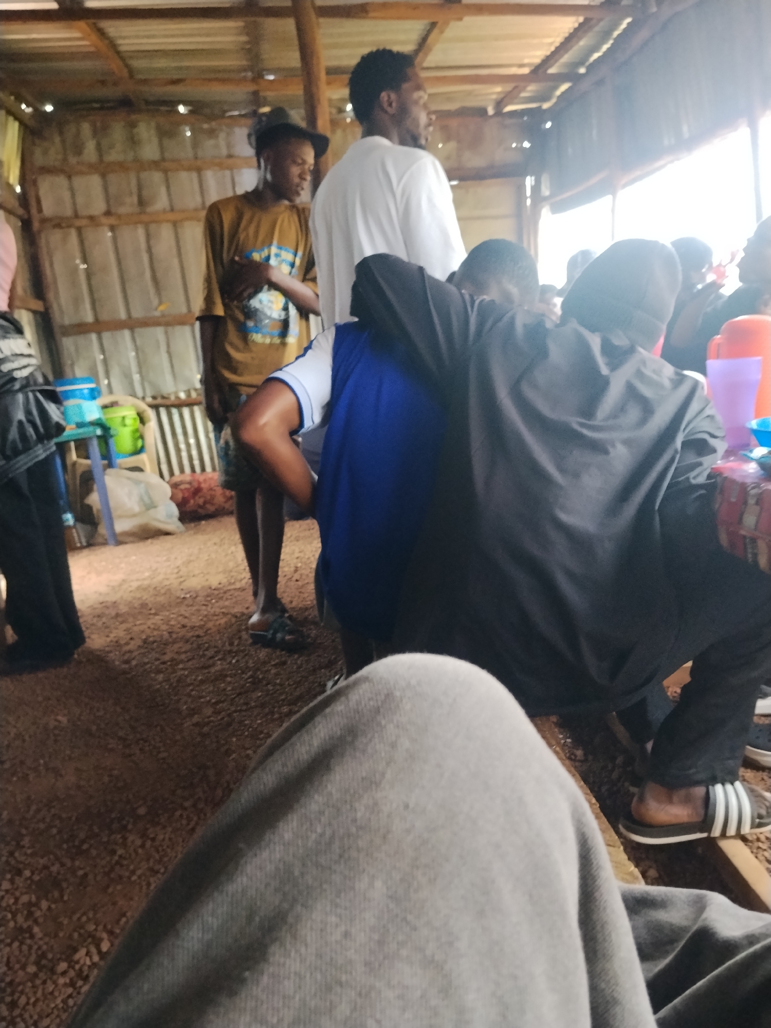 The image depicts a group of people gathered indoors, likely in a casual setting. The subjects include several individuals, some of whom are wearing head coverings and traditional African attire. The scene is set within a rustic structure with corrugated metal roofing supported by wooden beams. The walls appear to be made of corrugated metal as well, creating a simple and utilitarian atmosphere.
The people are engaged in what seems to be a social gathering, possibly a meal or a discussion. Some individuals are seated, while others are standing. The foreground is dominated by the legs of a person wearing gray pants, suggesting a first-person perspective or a close-up shot. In the background, there are various objects, including plastic containers, cups, and bags, suggesting provisions or supplies.
Notable details include the presence of a young boy in a brown t-shirt and a hat, looking down. Another person is wearing a blue t-shirt. The lighting suggests it might be daytime, possibly late morning or early afternoon, given the overall brightness. The environment appears to be an open-air or semi-open structure, typical of some informal gathering spaces in Nigeria. There are no clear indicators of specific emotions, but the gathering suggests a sense of community and shared activity. No visible text is present in the image. The ground is covered with small stones or gravel.