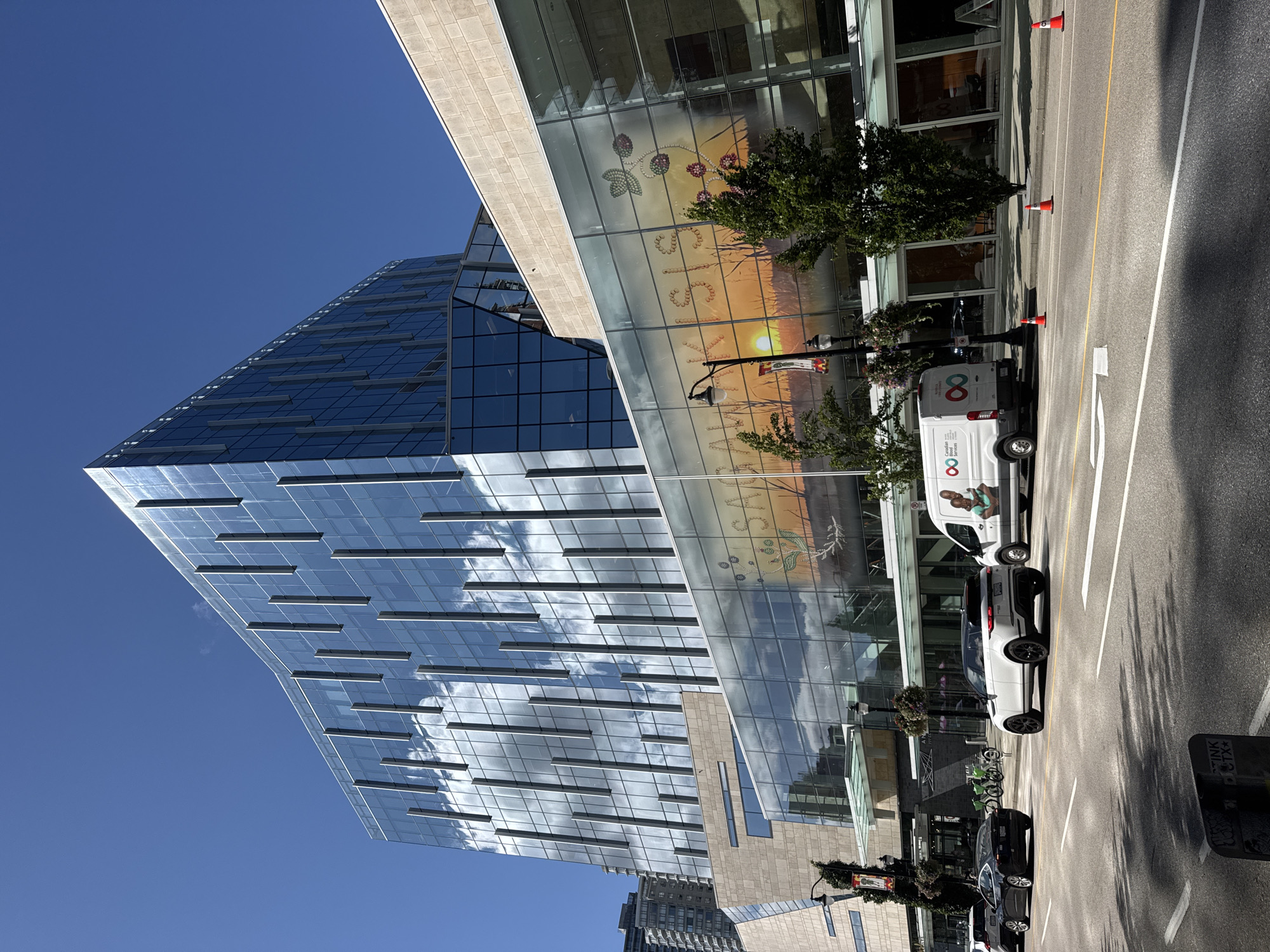 This image captures a street view in New Westminster, Canada, featuring a modern, glass-facaded building. The building's exterior reflects the bright blue sky and scattered clouds. A large graphic, featuring a sunset and floral designs, is visible on the lower section of the glass facade. Several vehicles, including a white SUV, a black sedan, and a white van with the logo "Canadian Services", are on the road. Orange traffic cones are placed on the street. Green bicycles are parked on the sidewalk. Streetlights and trees line the street. The overall environment suggests a sunny day in an urban setting.