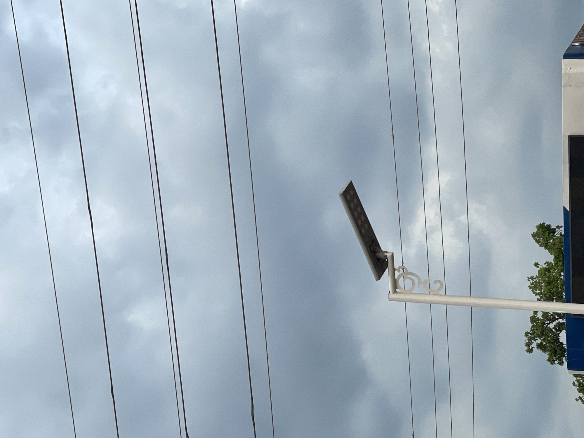 The image shows a cloudy sky in Akwanga, Nigeria. Electrical wires traverse the scene diagonally. A modern LED street light is mounted on a pole. A portion of a building with a dark window or panel is visible on the right, partially obscured by green foliage.