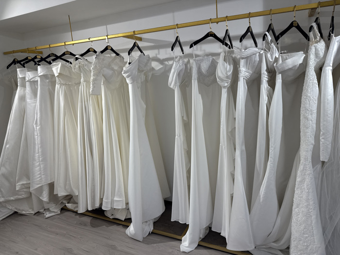 A row of elegant white wedding dresses hangs neatly on black hangers from a gold-colored rod. The dresses feature diverse styles, including strapless, off-the-shoulder, and long-sleeved designs. The fabric varies from smooth satin to delicate lace, creating a textured display of bridal attire. The dresses are all various lengths and some of them are covering the floor.