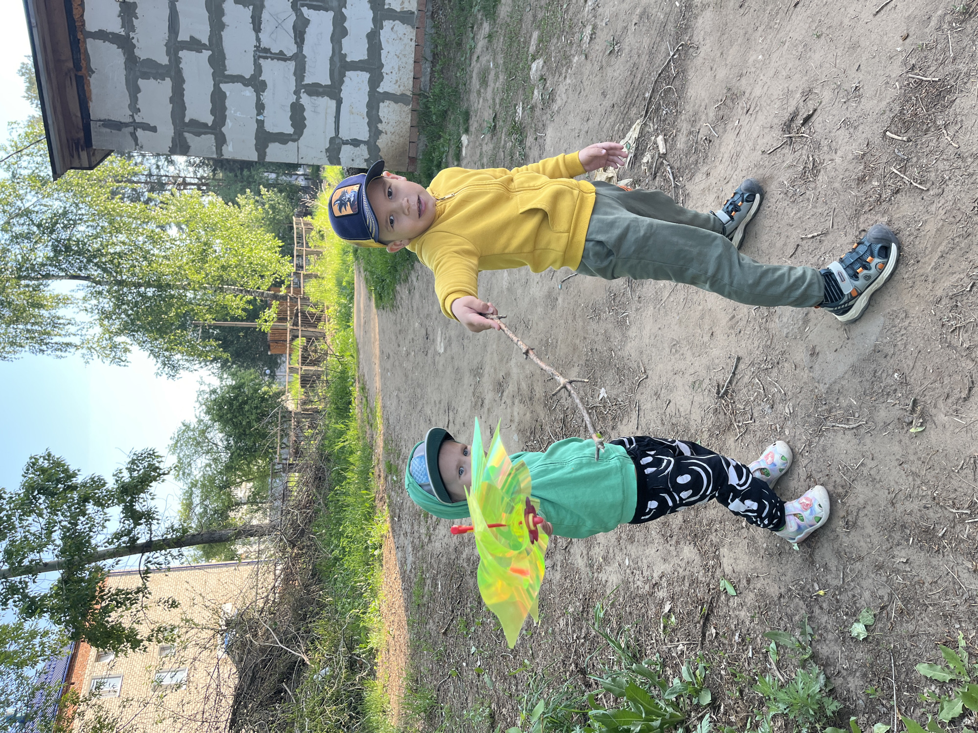 Two young boys stand on a dirt path. The boy on the left wears a green hooded sweatshirt, black and white patterned pants, and light-colored shoes, holding a yellow pinwheel on a stick. The boy on the right wears a yellow sweatshirt, green pants, a baseball cap, and dark-colored sneakers, holding a stick in his hand. The background shows a building constructed of concrete blocks, trees, and other buildings. The location is the city of Chaikovsky, Russia.
