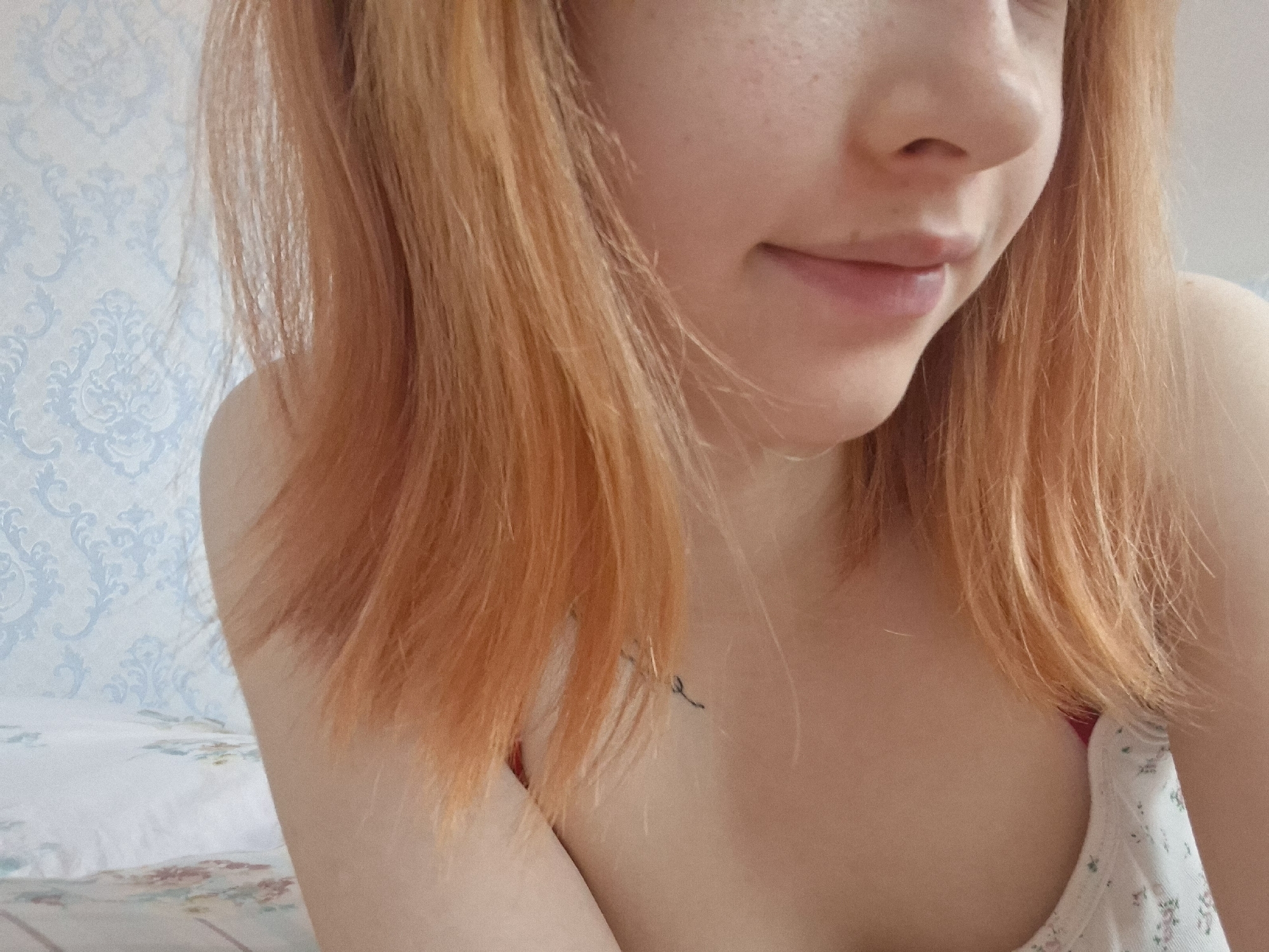 A close-up, medium shot shows the upper torso and face of a fair-skinned person with freckles and shoulder-length reddish-blonde hair. The person is partially smiling. They are wearing a white tank top with a floral pattern and thin red straps. A small, minimalist tattoo is visible on their upper chest/neck. The background is a light blue wallpaper with a repeating ornate pattern, and a bed with a floral patterned sheet. There is no specific information to indicate the location is in Chaikovsky, Russia.