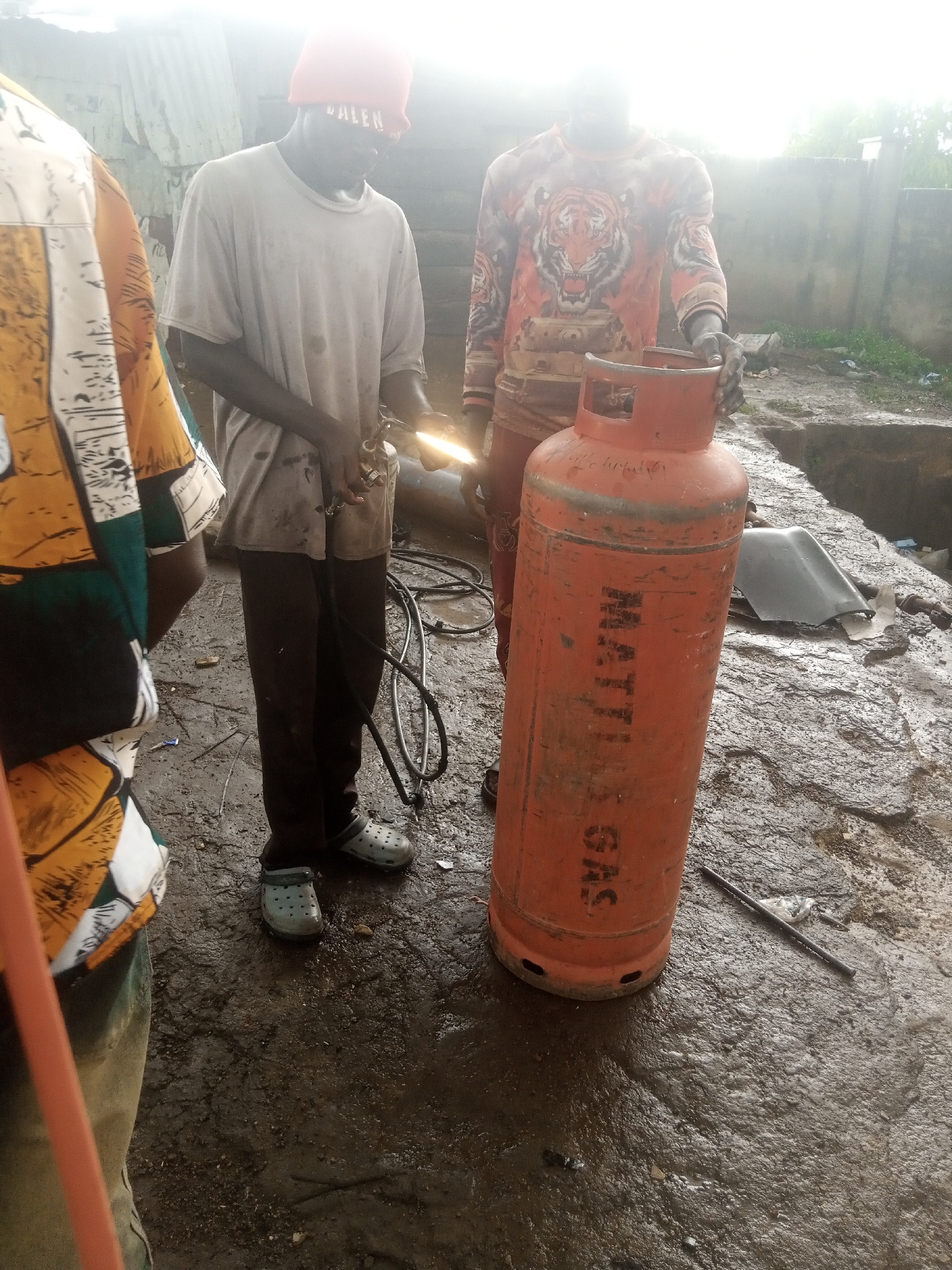 In Jos, Nigeria, three individuals are captured in an outdoor scene, likely a workshop or a yard, indicated by the wet, muddy ground and the corrugated metal roofing in the background. The lighting suggests it is daytime, possibly overcast or early morning/late afternoon due to the diffused light.
The central focus is a large orange gas cylinder standing upright. The text "MATTI GAS" is clearly visible vertically on its side in black lettering. To the left of the cylinder, a person wearing a white t-shirt and dark pants is holding a torch, appearing to be welding or working on something near the ground. This individual wears a red beanie with "VALEN" written on it in white. To the right of the gas cylinder, another person, wearing a long-sleeved shirt with a prominent tiger graphic in orange and black, stands with their hand resting on the top of the cylinder. A third person is partially visible on the far left, wearing a brightly colored shirt with a yellow, white, green, and black abstract pattern.
The ground is wet and reflective, with puddles and debris scattered around, suggesting recent rain or a generally damp environment. The atmosphere appears functional and perhaps a bit gritty, typical of a working area. The subjects' actions suggest a repair or maintenance activity involving the gas cylinder or related equipment.