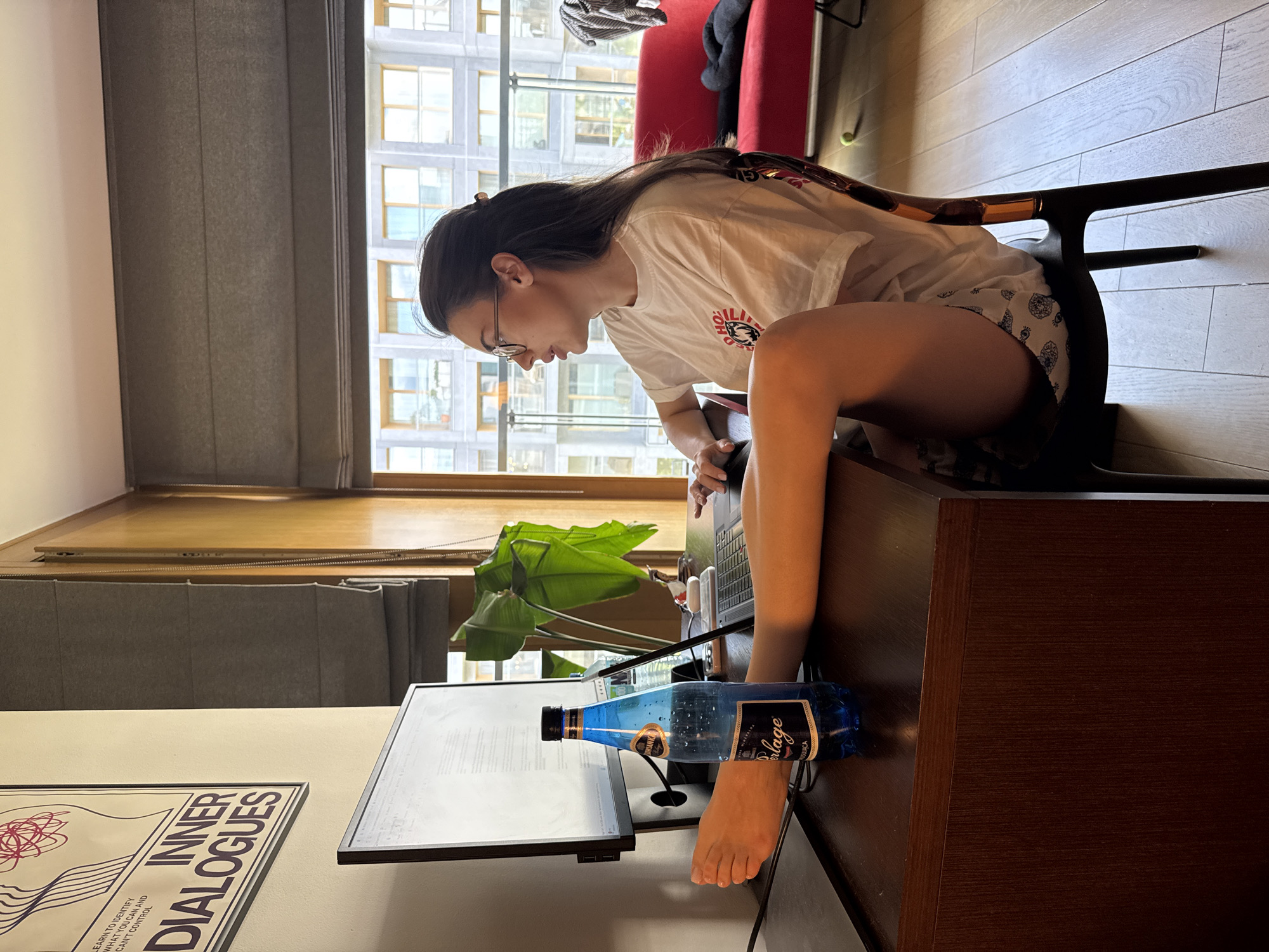 A woman is sitting at a desk, working on a laptop. Her left foot is resting on a bottle of water on the desk. The desk is in a room with a large window. A red chair is visible in the background. A poster for "INNER DIALOGUES" hangs on the wall.