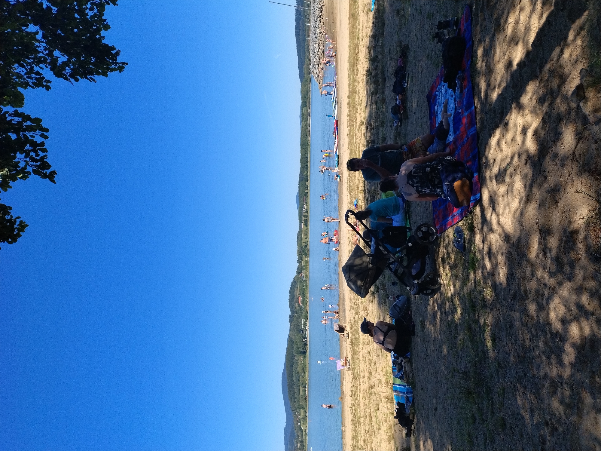 This is a photograph taken on a sunny day at the Radslav water reservoir in the Czech Republic. The scene depicts a sandy beach with several people enjoying a day by the water. In the foreground, there are people relaxing on beach towels and blankets, with a stroller nearby. Further in the background, many people are in the water, swimming and playing. The water is calm and blue, with a tree-covered hilly landscape in the distance. The sky is clear and a vibrant blue.