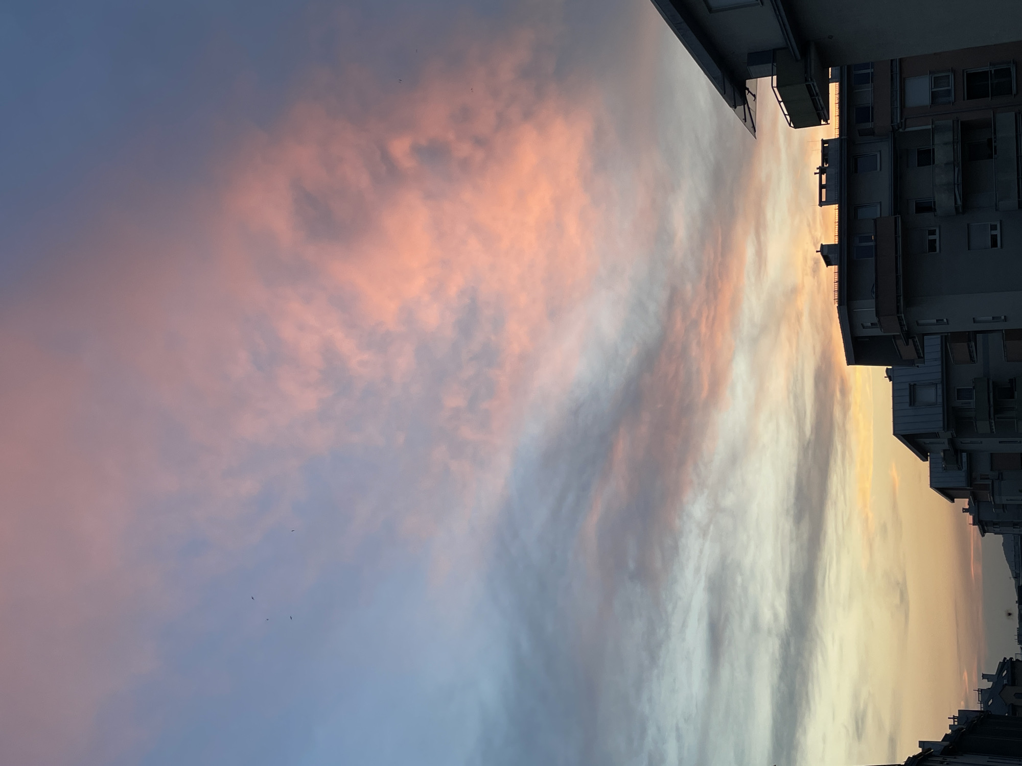The image shows a city skyline at sunset. The sky is filled with pink and orange clouds, with some areas of blue. There are several buildings visible in the foreground, including apartment buildings with balconies. The overall impression is one of a peaceful evening in an urban setting.
