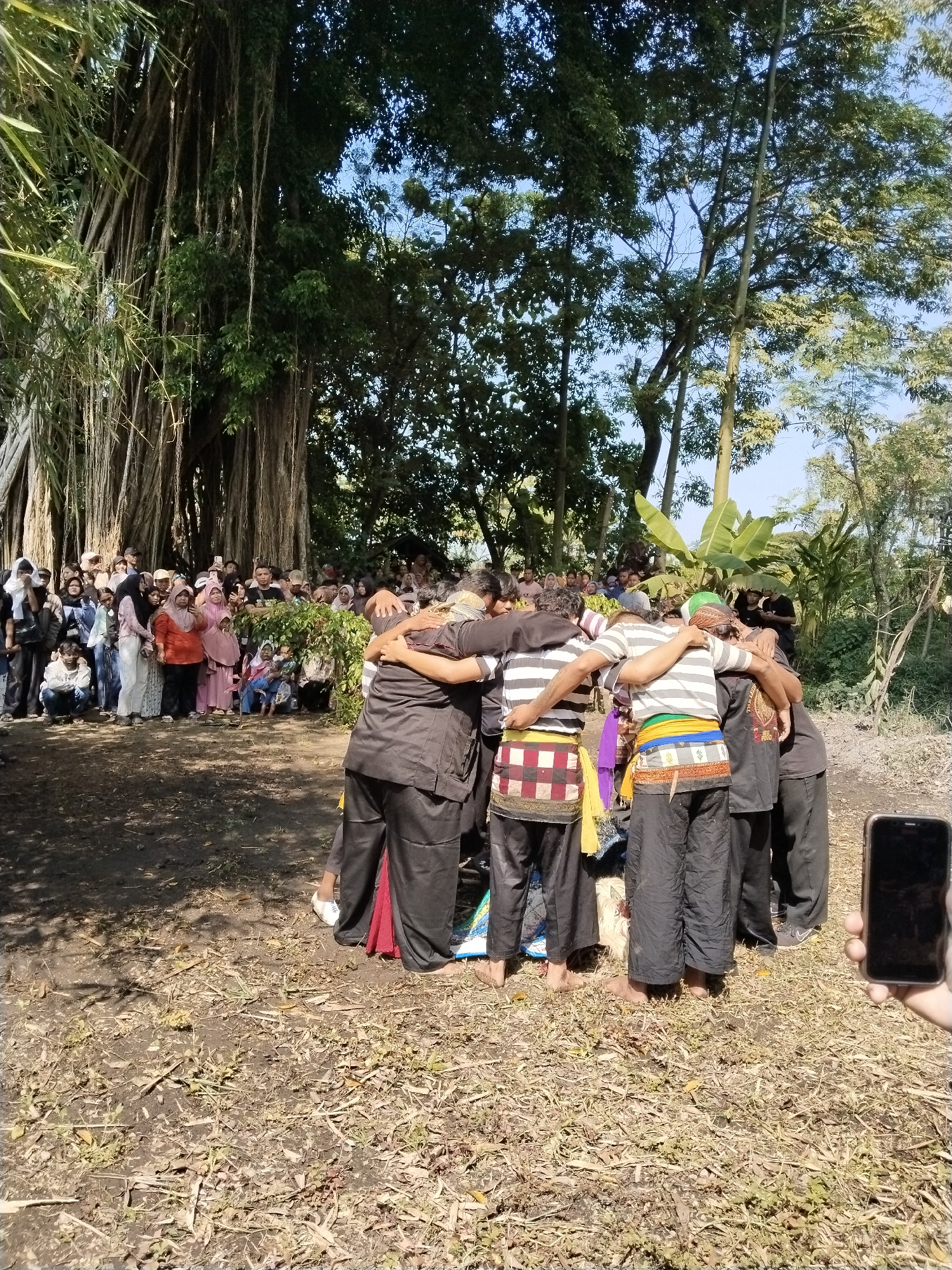A group of people, predominantly women, some wearing head coverings, stand in the background, observing a circle of men, tightly interlocked with their arms around each other’s shoulders. The men are dressed in dark, traditional clothing and patterned wraps around their waists. The event appears to be taking place outdoors in a semi-rural location with trees and foliage present. A hand holding a smartphone is visible in the foreground, suggesting someone is capturing the scene on camera. The location may be Gebang, Indonesia.