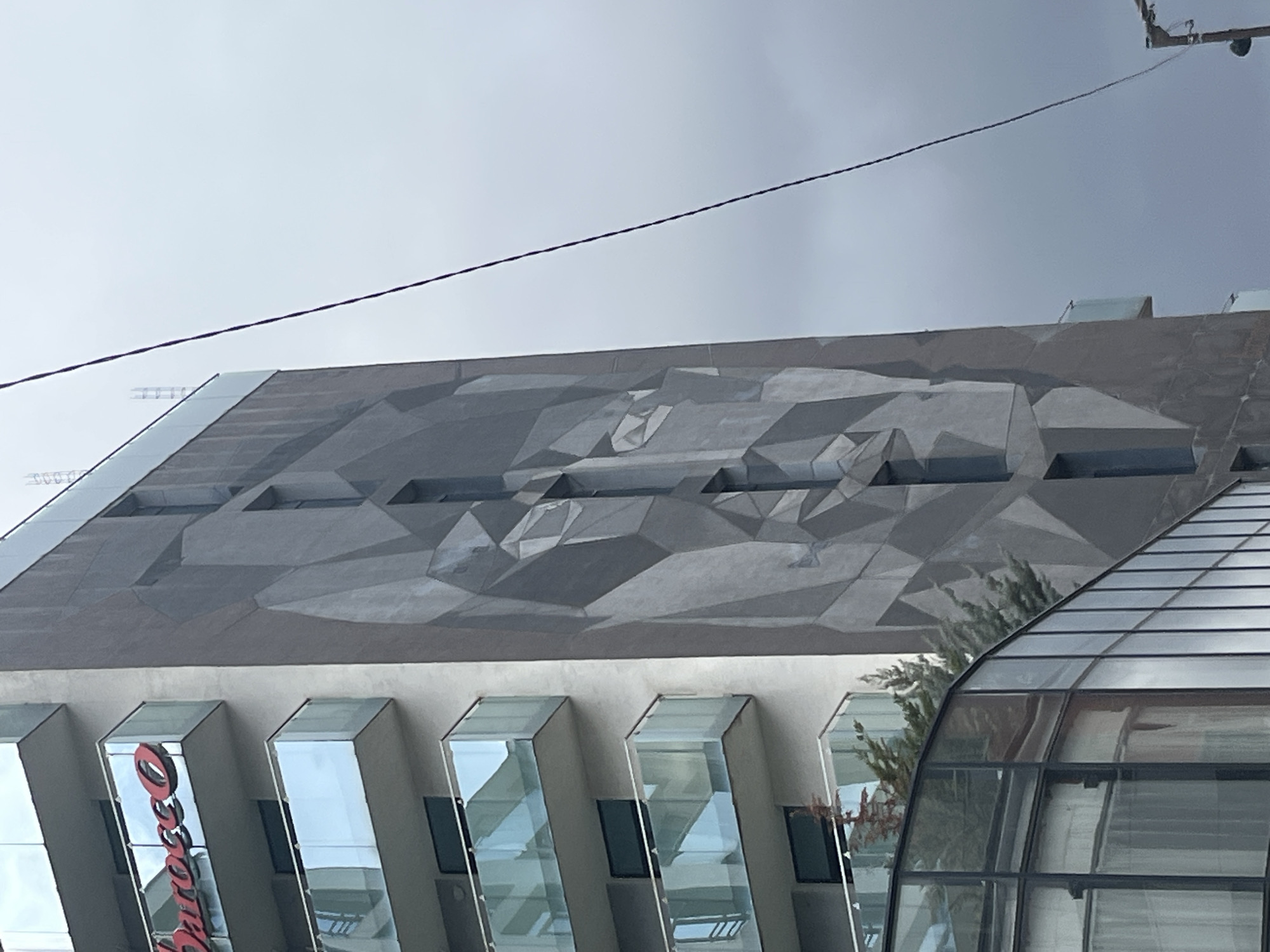 The image shows a section of a modern building in Sofia, Bulgaria, with a distinctive geometric mural on the upper portion of the facade. Below the mural, the building features a series of vertical glass windows that reflect the surroundings. A curved glass structure is visible to the right. A power line crosses the upper portion of the frame against a cloudy sky. A portion of the red and white "Oscars" branding is visible in the lower left, indicating it could be the Oscars Cinema in Sofia.