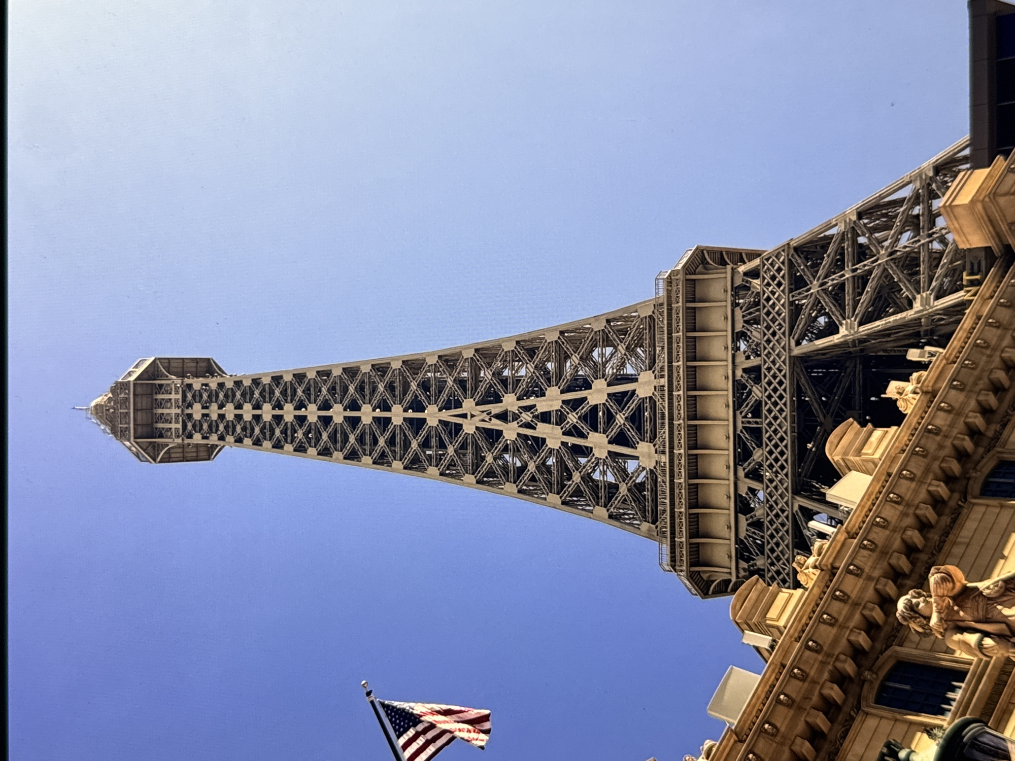 The image shows a low-angle view of the Eiffel Tower at the Paris Las Vegas Hotel & Casino on the Las Vegas Strip in Paradise, Nevada, United States of America, under a clear blue sky. A portion of an ornate building with architectural details and statues is visible in the foreground, directly beneath the tower. An American flag is prominently displayed on a flagpole to the left of the tower, partially obscuring the clear blue sky. The Eiffel Tower is a replica of the original Eiffel Tower in Paris, France, and is a landmark attraction in Las Vegas.