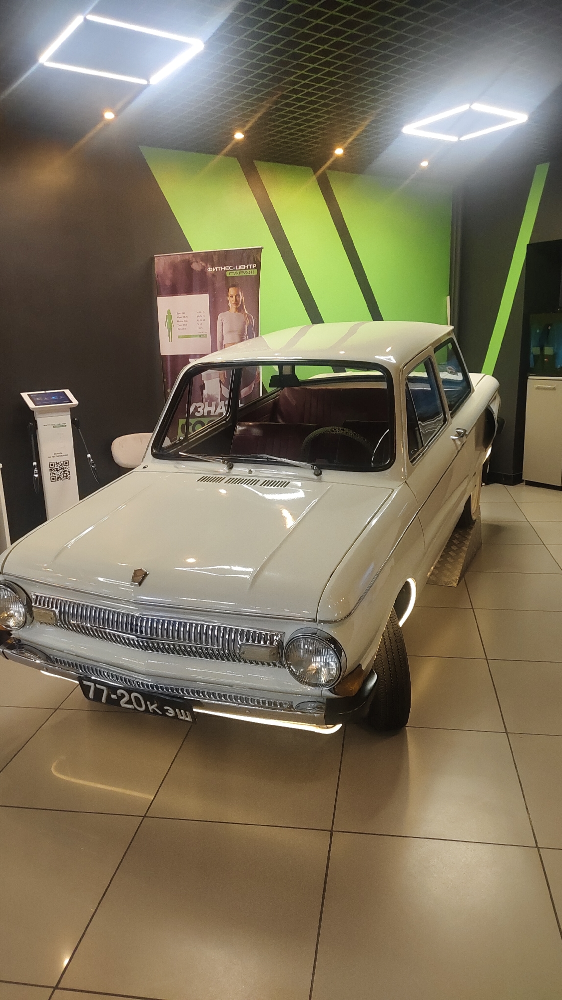 A white, vintage ZAZ-966 Zaporozhets car is displayed indoors, likely in a public space in Krasnoyarsk, Russia. The car has a license plate reading "77-20кэш". The interior is visible through the windows, featuring maroon-colored seats. The car is positioned on a metal ramp.
Behind the car is a fitness center advertisement featuring a woman, and a black wall with green geometric accents. To the left is a white electronic device on a stand. The floor is tiled in a neutral tone and the ceiling has modern, square lighting fixtures. The environment appears clean and well-lit.