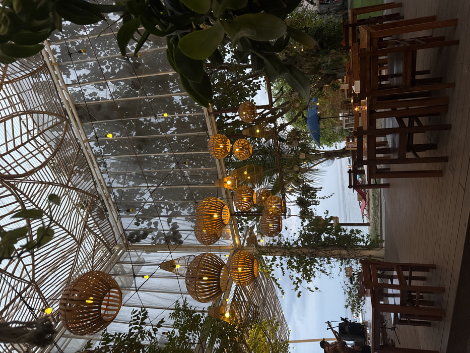 The image captures an outdoor dining or lounge area situated directly on a beachfront in Ped, Indonesia, during dusk or early evening. The overhead structure features an intricate wooden or bamboo lattice ceiling, transitioning into transparent panels adorned with string lights. Numerous woven rattan or bamboo pendant lamps, varying in size, are suspended from the ceiling, emitting a warm, orange-yellow illumination. Green foliage, including hanging plants, is integrated into the environment. The floor consists of dark wooden decking with multiple unoccupied wooden tables and chairs, some holding small lit candles. To the left, two individuals are present near the beach; one is playing an acoustic guitar next to a dark speaker. Beyond the establishment, a sandy beach is visible with red lounge chairs and a blue umbrella, leading to a calm body of water under an overcast sky.