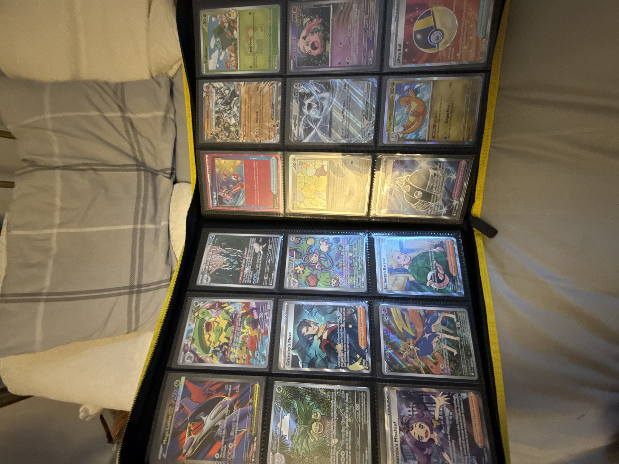The image shows a close-up, eye-level, full shot of a black zippered binder filled with Pokémon cards. The binder is laid open on a bed with a grey and white plaid comforter. The binder has yellow trim around the zipper. Inside the binder, the cards are arranged in a grid of three columns and multiple rows. The top row displays cards featuring Mega Latias, Ludicolo, and Clawitzer. The second row shows Max Rod, Genesect, and Bulbasaur. The third row features Exeggutor, Grimsley's Move, and Ambipom. The fourth row has Acerola's Mischief, Zacian, and Lt. Surge's Bargain. The fifth row displays Vikavolt Helm, Dragonite, and Ultra Ball. The lighting is dim, and there is no visible indication of the city or location.
