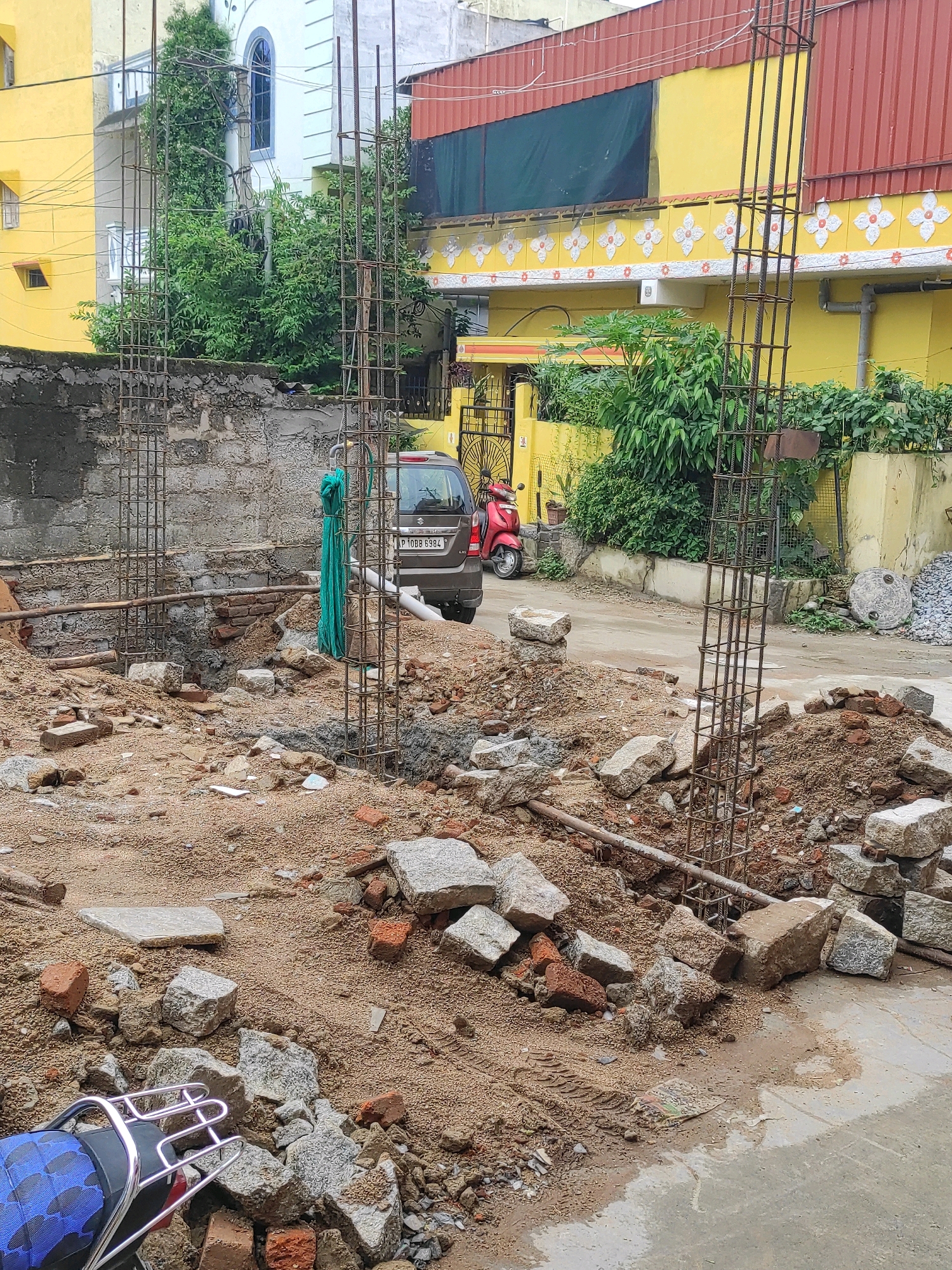 The image depicts an outdoor scene of ongoing construction in what appears to be a residential area of Hyderabad, India. In the foreground, loose soil, construction debris including bricks and stones, and a section of wet concrete are visible. Two rebar cages, forming structural columns, are prominently featured, indicating the foundation work for a building. A green tarpaulin is draped over a horizontal pipe.
In the mid-ground, a gray car with the license plate P 10BB 6984 is parked on a paved surface. Behind the car, a red scooter is also present. A roughly constructed concrete wall runs across the middle ground, with overgrown green foliage visible behind it.
The background reveals residential buildings. To the left, a yellow building with white trim and a tall arched window is partially visible. To the right, a bright yellow building with decorative white floral patterns along its facade and a red corrugated metal roof is prominent. The environment appears to be a street or alleyway within a city. The overall impression is one of a developing or under-renovation urban landscape.