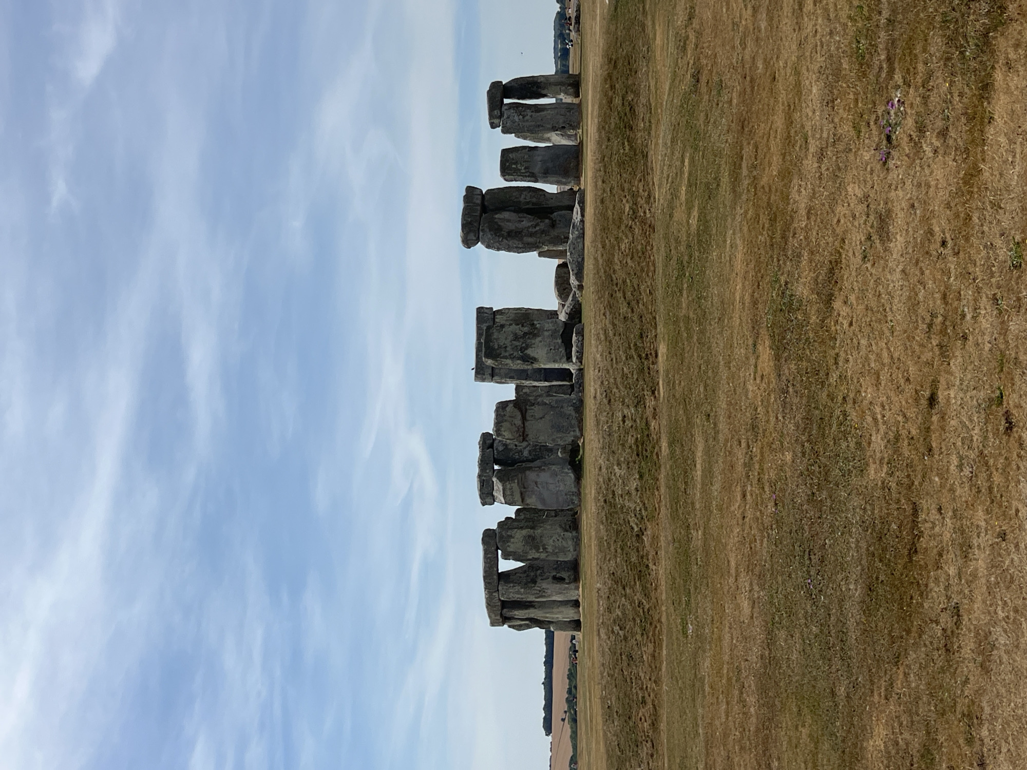 The image depicts Stonehenge, a prehistoric monument located near Amesbury, United Kingdom. The monument consists of a circle of large standing stones, some of which are topped with horizontal lintels. The stones are grey and weathered, with rough textures. They are arranged in a circular formation on a flat, grassy plain. The grass in the foreground and middle ground is dry and brown, indicating a sunny or arid climate. In the background, there are rolling green hills and a few scattered trees. The sky is a pale blue with wispy white clouds. Several tourists are visible in the distance, walking around the monument. The overall scene is one of historical significance and natural beauty.