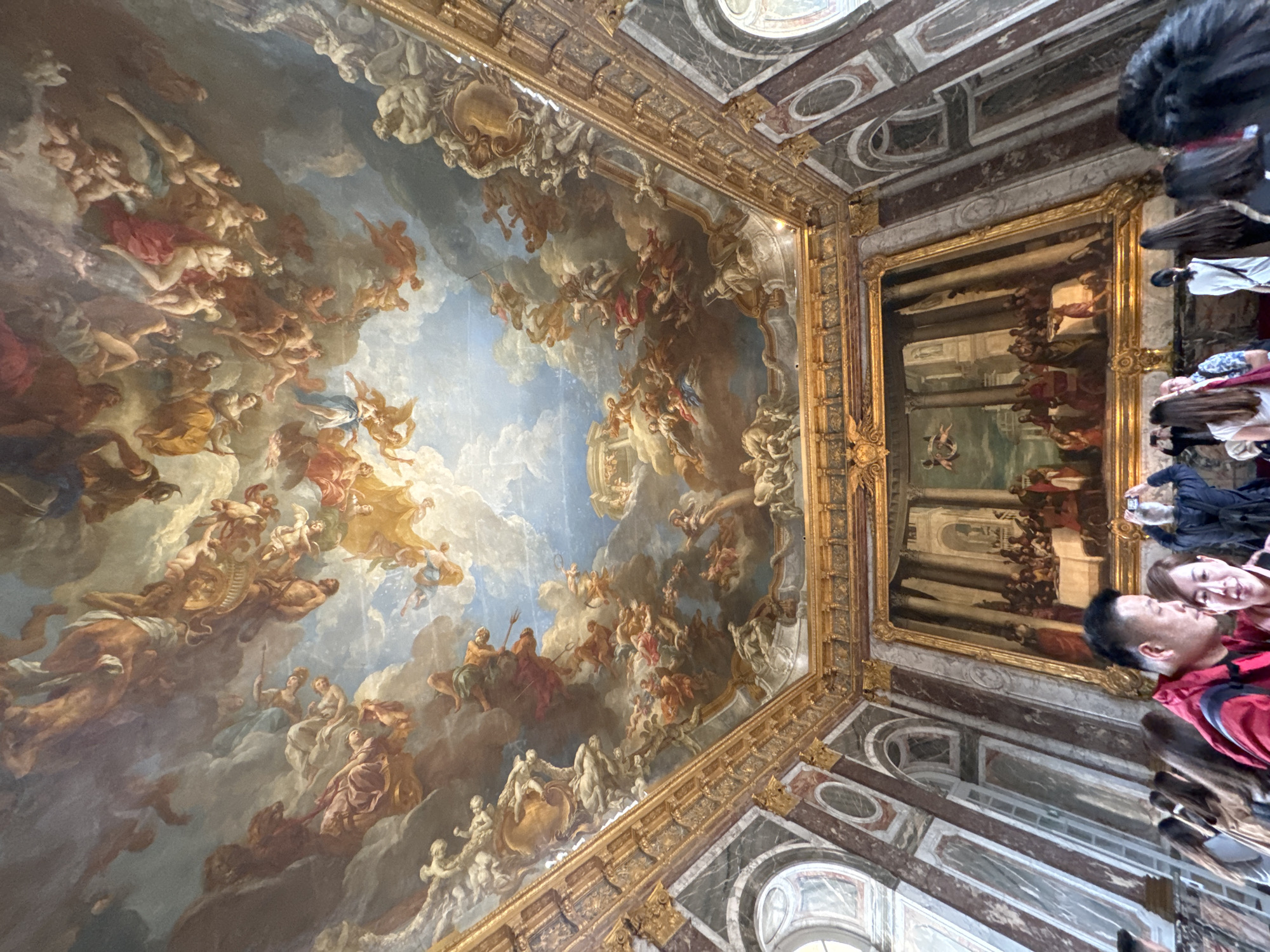 The image captures the ornate ceiling and wall decorations within the Palace of Versailles, located in Versailles, France. The ceiling features a large-scale painting depicting an allegorical scene with numerous figures in varying poses, set against a backdrop of clouds and sky. The painting is framed by an elaborate gold trim. Adjacent to the ceiling painting, a large framed painting hangs on the wall, portraying a formal gathering or banquet scene with many people in classical attire. The wall is clad in marble with geometric patterns and framed arched windows. Several people are visible on the right side of the frame, presumably tourists looking up at the artwork. One person is visible in the front right of the frame wearing a red jacket.
