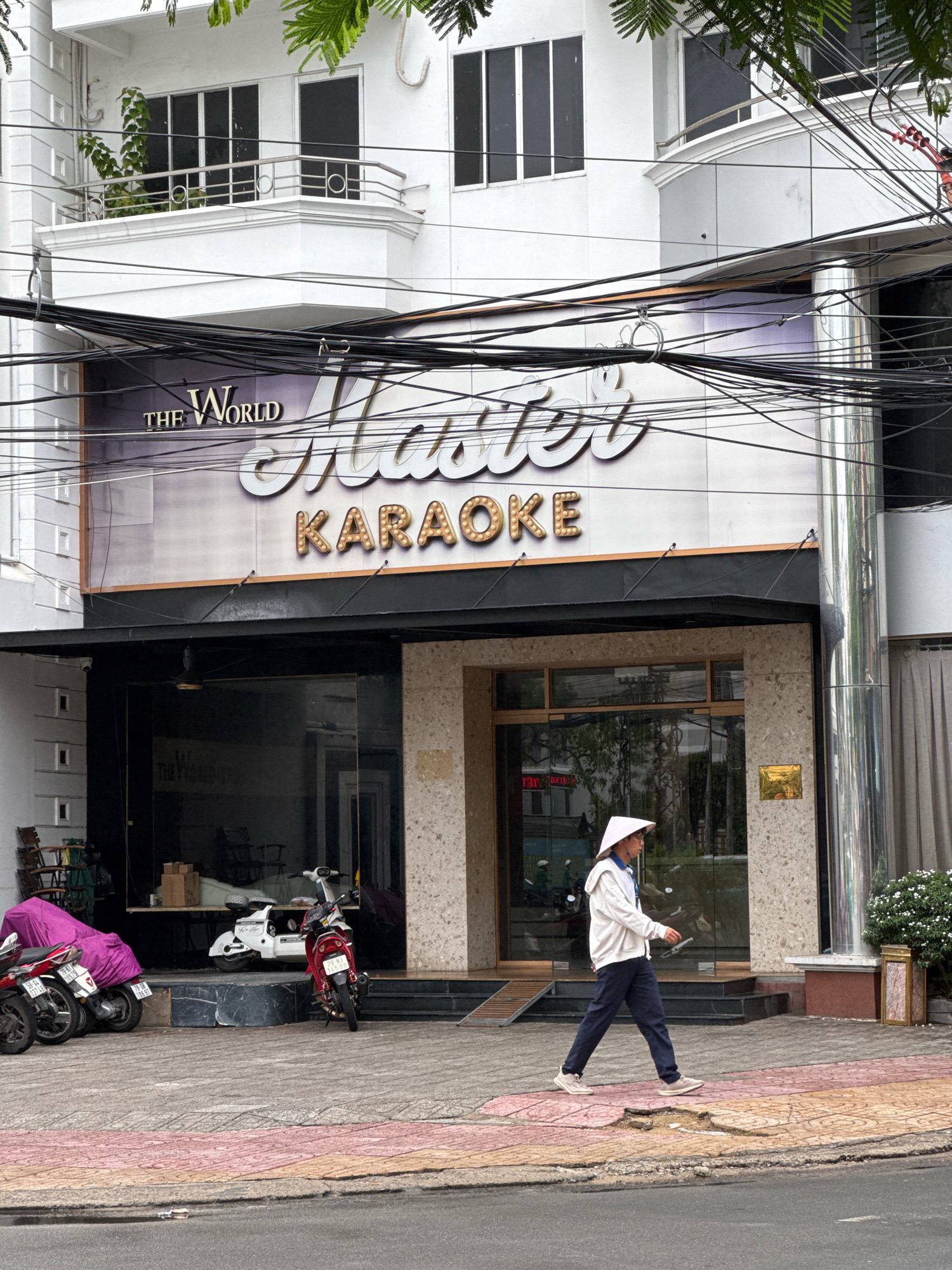The image shows the exterior of a building in Nha Trang, Vietnam, with a sign that reads "The World Master Karaoke". A person wearing a conical hat and casual clothing is walking on the sidewalk in front of the building. Several motorcycles are parked on the side of the street. The building appears to be a commercial establishment. The overall scene is typical of a street in a Vietnamese city.