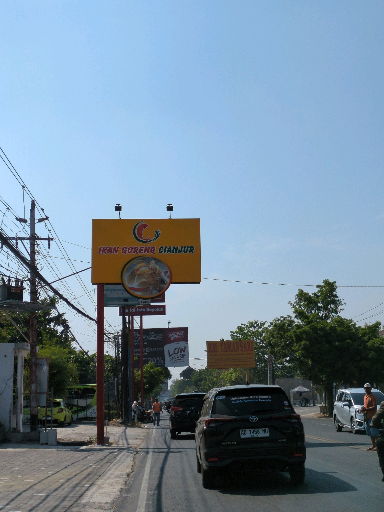 The image depicts a street scene in Colomadu, Indonesia, on a clear, sunny day. The foreground features a road with several vehicles, including a black Toyota Avanza with the license plate "AD 1958 MU" and a silver minivan. A person in an orange shirt is standing next to the silver minivan. On the left side of the road, a yellow car is parked.
Dominating the upper portion of the image is a large yellow billboard advertising "IKAN GORENG CIANJUR" (Fried Fish Cianjur), featuring an image of fried fish. Below this, other billboards are visible, including one that appears to advertise "RM TAMANSARI". A utility pole with numerous wires is prominently situated on the left side of the image.
In the mid-ground, several individuals are walking along the side of the road, near some parked motorcycles. Lush green trees line the right side of the street, and beyond them, some structures and tents are visible. The overall environment suggests a commercial or roadside area with active traffic.
