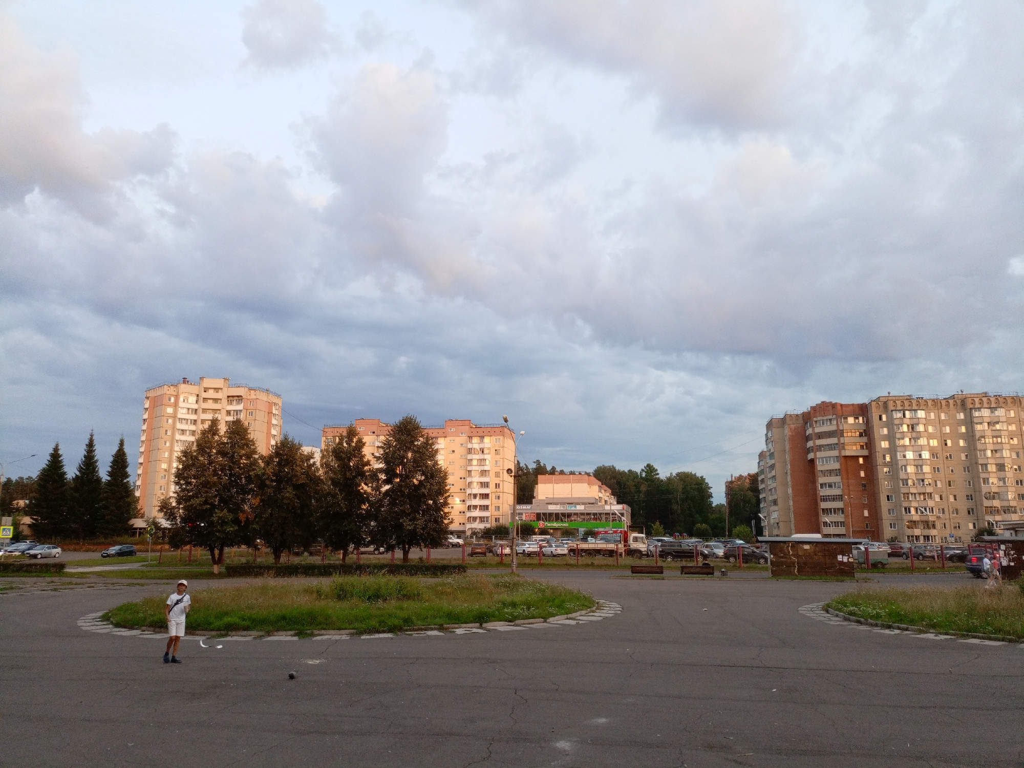 A full shot captures a broad, paved area with a circular grassy median in the foreground, on a cloudy day in Zheleznogorsk, Russia. A young person wearing a white t-shirt, white shorts, a white cap, and dark shoes stands in the paved area on the left, facing the camera, with a small white object on the ground in front of them. The area is surrounded by a line of trees and a multi-story apartment buildings on the background. The buildings are light-colored with numerous windows and balconies. A sign with Russian text is visible on one of the buildings. Several cars are parked in front of the buildings. The sky is filled with large, gray clouds, some of which are tinged with pink from the setting sun. The overall scene depicts a typical urban environment on a late afternoon.