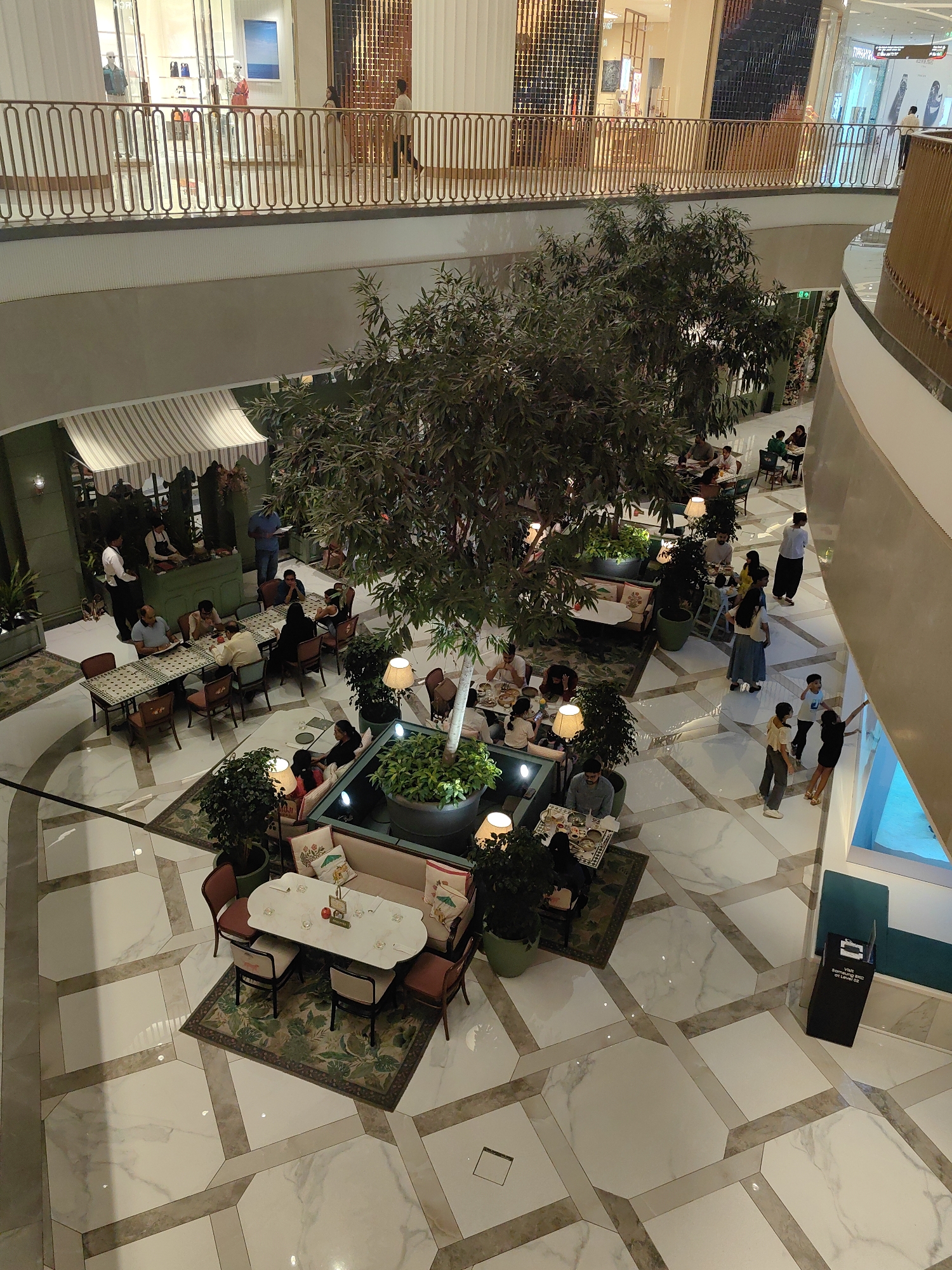 This is an overhead shot of the Palladium Mall in Lower Parel, Mumbai, India. The scene captures the interior of the mall, focusing on a restaurant or cafe area on the ground floor, with a walkway and shops visible on the level above.
The foreground shows a spacious, open-plan area with a marble-tiled floor featuring a geometric pattern. In the center of the shot, a large, leafy tree stands prominently, acting as a focal point. Surrounding the tree are several tables and seating arrangements for patrons.
To the left of the tree, there are multiple dining tables with chairs, occupied by individuals and groups engaged in conversations and meals. A long table with several people seated is visible, with two servers attending to them. Further left, an awning covers a section of the seating area.
To the right of the tree, a section of a contemporary seating area with plush sofas and cushions is visible, with people relaxing. There are also additional tables and chairs in this area. Decorative lamps are strategically placed around the seating areas.
On the upper level, a walkway with a decorative railing overlooks the lower floor. Several shops with glass storefronts are visible, showcasing displays and mannequins. People are seen walking along the walkway.
The overall atmosphere is one of a busy, upscale mall with dining facilities. The lighting is bright and even, suggesting daytime or well-lit interior conditions. The color palette is dominated by neutral tones, greens from the plants, and the warm tones of the wood and railings.