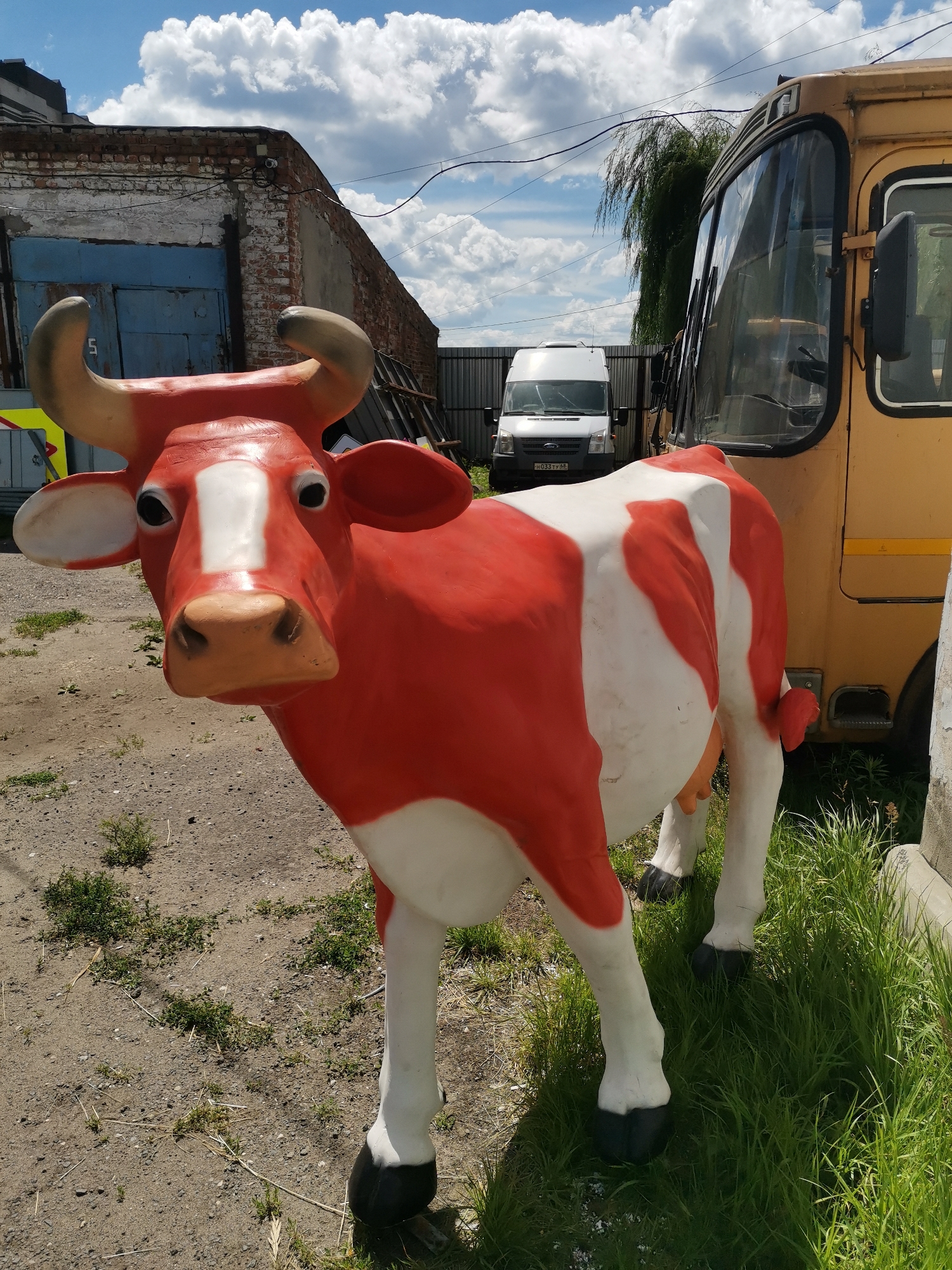 A red and white cow statue stands in an outdoor industrial area. The statue is life-sized and appears to be made of fiberglass or plastic. It has black hooves, prominent horns, and a realistic facial expression. The background features a yellow bus, a white van, a brick building with blue doors, a metal fence, and a cloudy blue sky. The ground is covered with gravel and sparse vegetation. This scene is located in the city of Rasskazovo, Russia, likely an industrial or agricultural facility. There are no people visible in the image, and no actions are taking place.