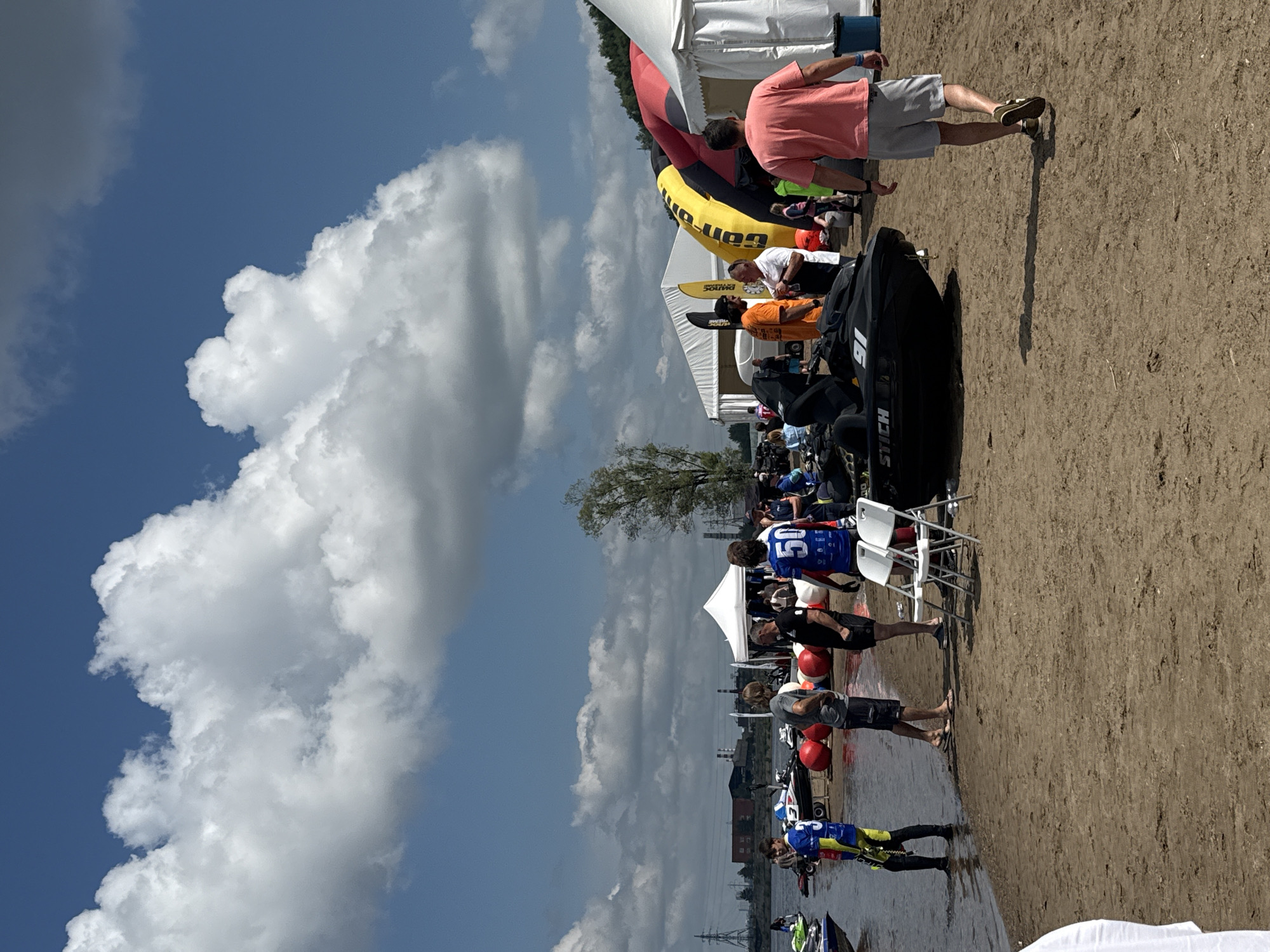 The image depicts an outdoor event on a sandy shore, likely near a body of water. The sky is blue with scattered white clouds. In the background, there are several white tents and inflatable structures, one of which is yellow and red. Industrial structures and power lines are visible in the far distance, suggesting a location near an industrial area within the city of Perm, Russia.
In the foreground, people are present on the sand. Some are standing and conversing, while others are walking. Several jet skis are parked on the shore. One black jet ski prominently displays the word "STICH" on its side. There are also large red inflatable buoys on the sand. Two white folding chairs are set up near the water.
Individuals in the image are dressed in casual attire. Some are wearing athletic-style jerseys with numbers, such as "3" and "50". One person in a pink t-shirt and grey shorts is walking away from the camera. Another person in a blue and yellow jersey is standing in the shallow water.
