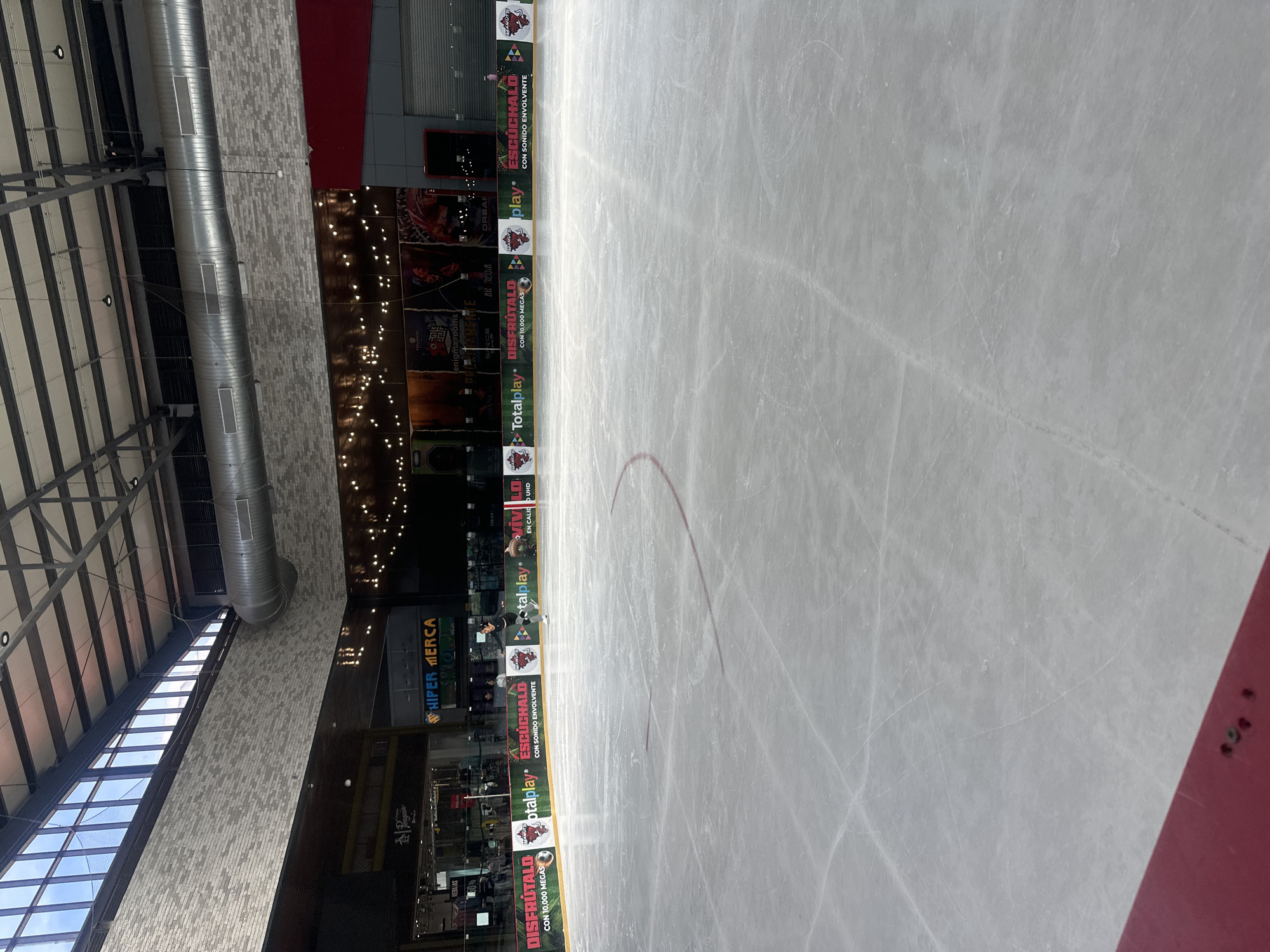 The image shows an indoor ice rink in Cholula, Mexico, likely part of a commercial establishment given the retail store facades visible in the background. The ice surface is marked with red lines and numerous skate marks. A single figure, dressed in dark athletic wear, is actively ice skating. The background features a large, modern ceiling with exposed ductwork and lighting. Along the perimeter of the rink, a colorful banner displays various logos and text, including "DISFRÚTALO CON 10,000 MEGAs" and "Totalplay," suggesting sponsorship or advertising. Storefronts with signage like "HIPER MERCA" and "La Pagua" are visible behind the banner, indicating the rink is located within or adjacent to a shopping area. The lighting appears to be a combination of overhead ambient light and decorative string lights. The overall scene depicts a recreational activity within an urban commercial setting.