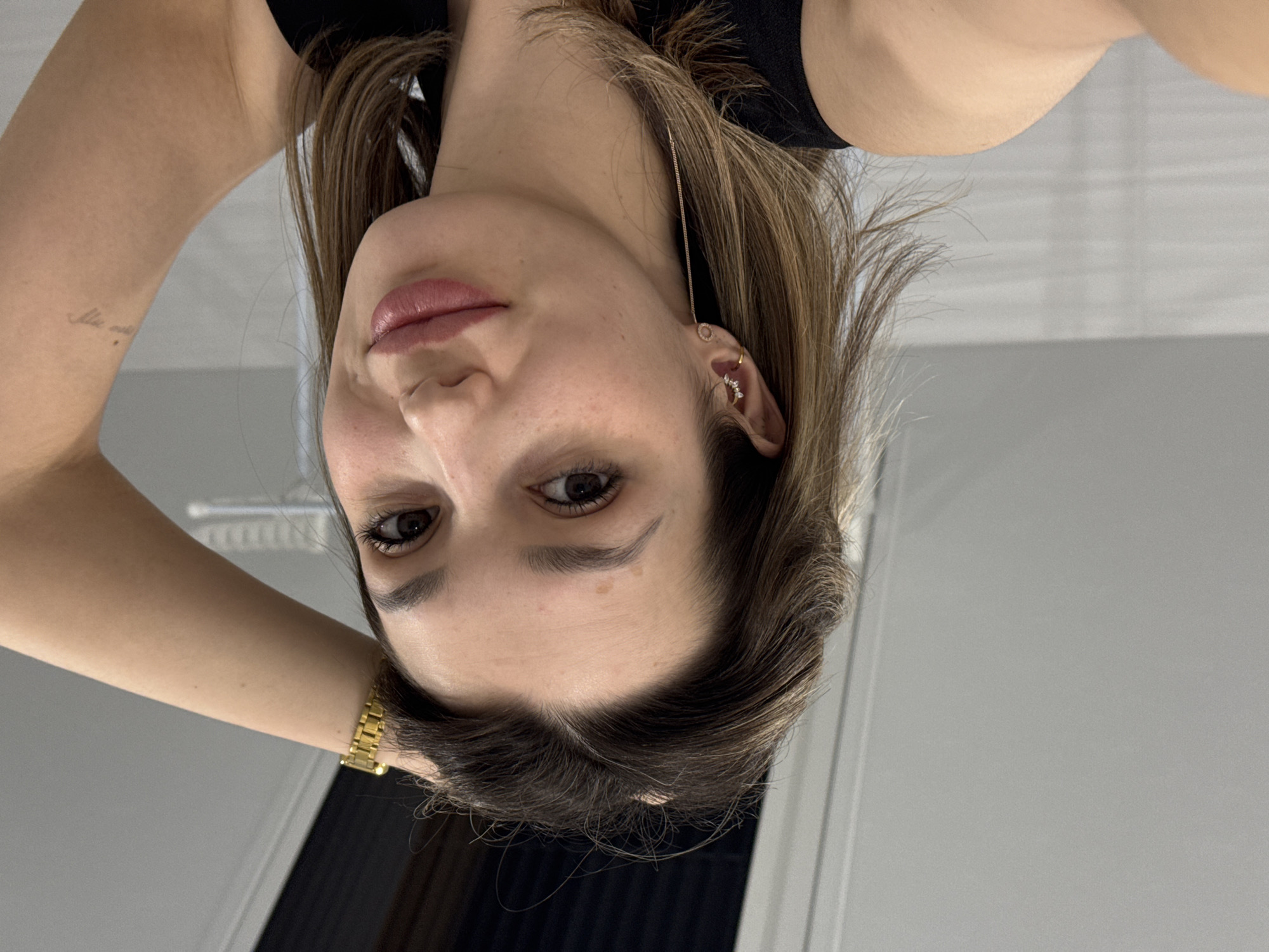 The image shows a woman with brown hair and brown eyes. She is wearing a black shirt and a gold watch on her left wrist. She has a small tattoo on her left arm. Her right ear has multiple piercings. The background is a white wall. The location is unknown.