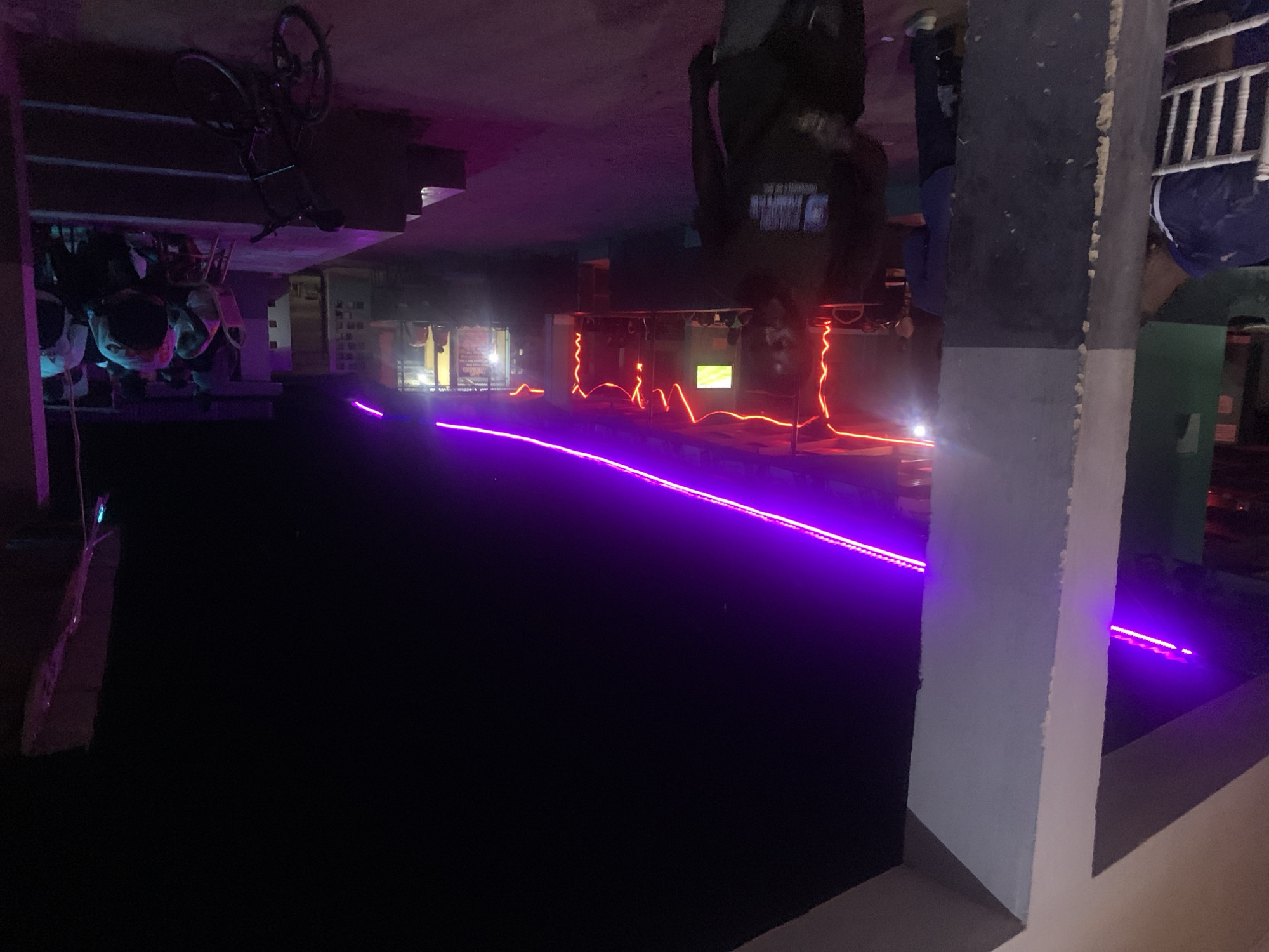 The image depicts a night scene in New Stadium, Nigeria. The photo is taken under the cover of a roof, which obscures some overhead elements. Under the roof, the area is lit by a line of bright purple LED strip lights that extend across the scene. To the left, some bicycles are either mounted to the roof or otherwise suspended, and several chairs are stacked. The primary focus of the scene is a building in the background, which has an open frontage with red neon lighting outlining its structure. The building appears to be a bar or restaurant. There is a vertical concrete pillar in the right foreground with a white top and gray bottom section. The photo also suggests the presence of individuals within the bar area, as seen in blurred silhouettes, although the details are not clear. The location appears to be urban and modern due to the lighting.