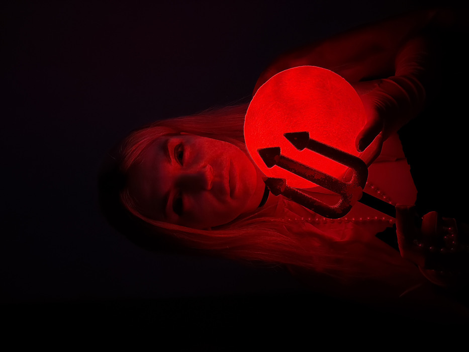 A woman with long blonde hair illuminated by red light holds a round, glowing red prop with a trident attached. She wears a black choker and possibly a beaded necklace. The dark background emphasizes the dramatic red lighting, casting the woman and prop in a sinister, theatrical light. Her expression is difficult to discern due to the intense red glow, but her face is partially visible.