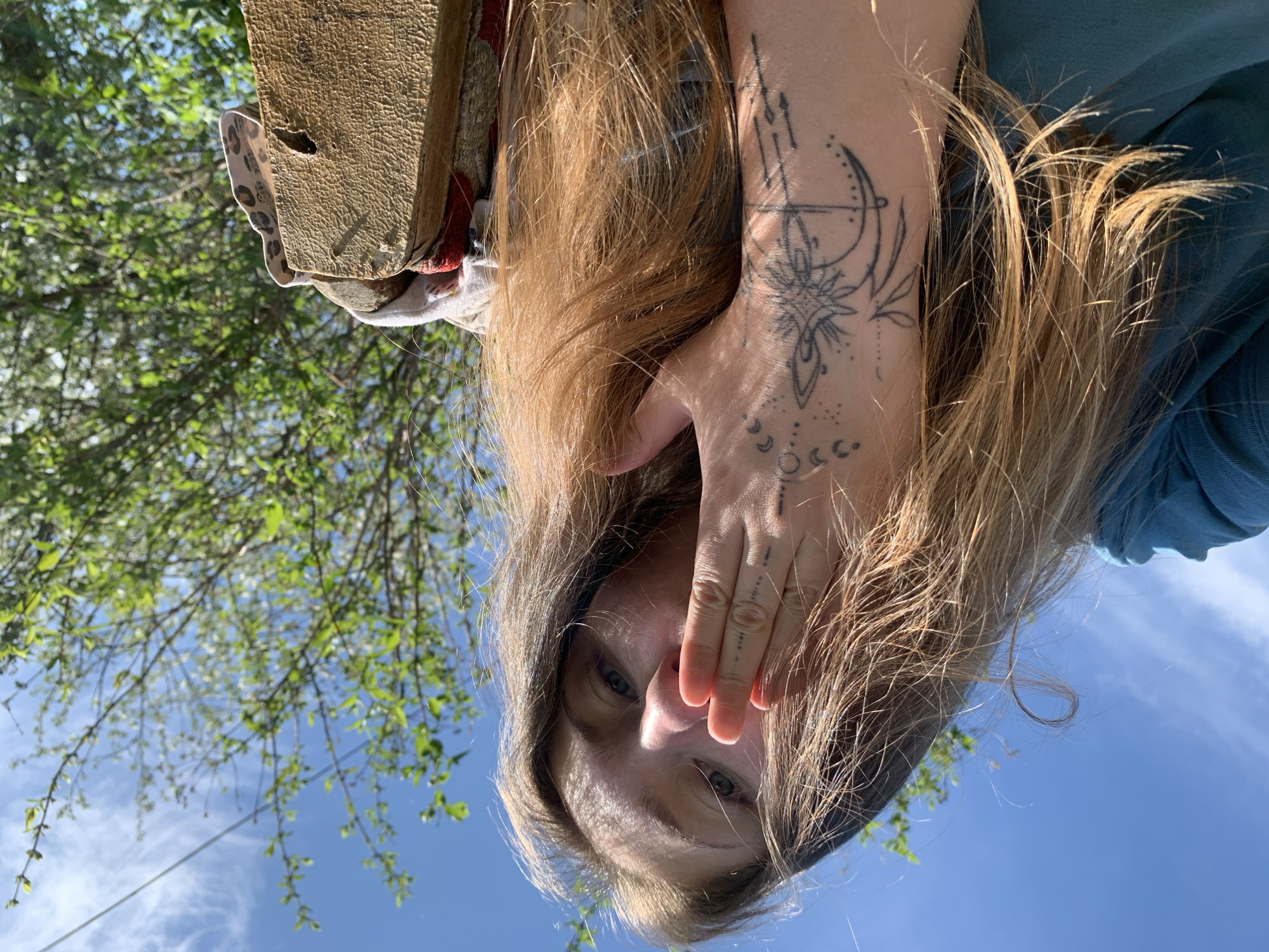 A woman with long, light brown hair is positioned with her face toward the camera, slightly obscured by her hair and her right hand, which covers her mouth. The woman's eyes are visible. Her right arm, exposed from the elbow down, displays intricate tattoo designs, including celestial motifs. The woman wears a blue shirt or blouse. The background consists of a blue sky with scattered clouds and green tree branches with leaves. A piece of wood is visible, possibly held by the woman. The location appears to be outdoors during daylight. The city of Zasechnyi, Russia, is not discernible from the visual elements in the image.