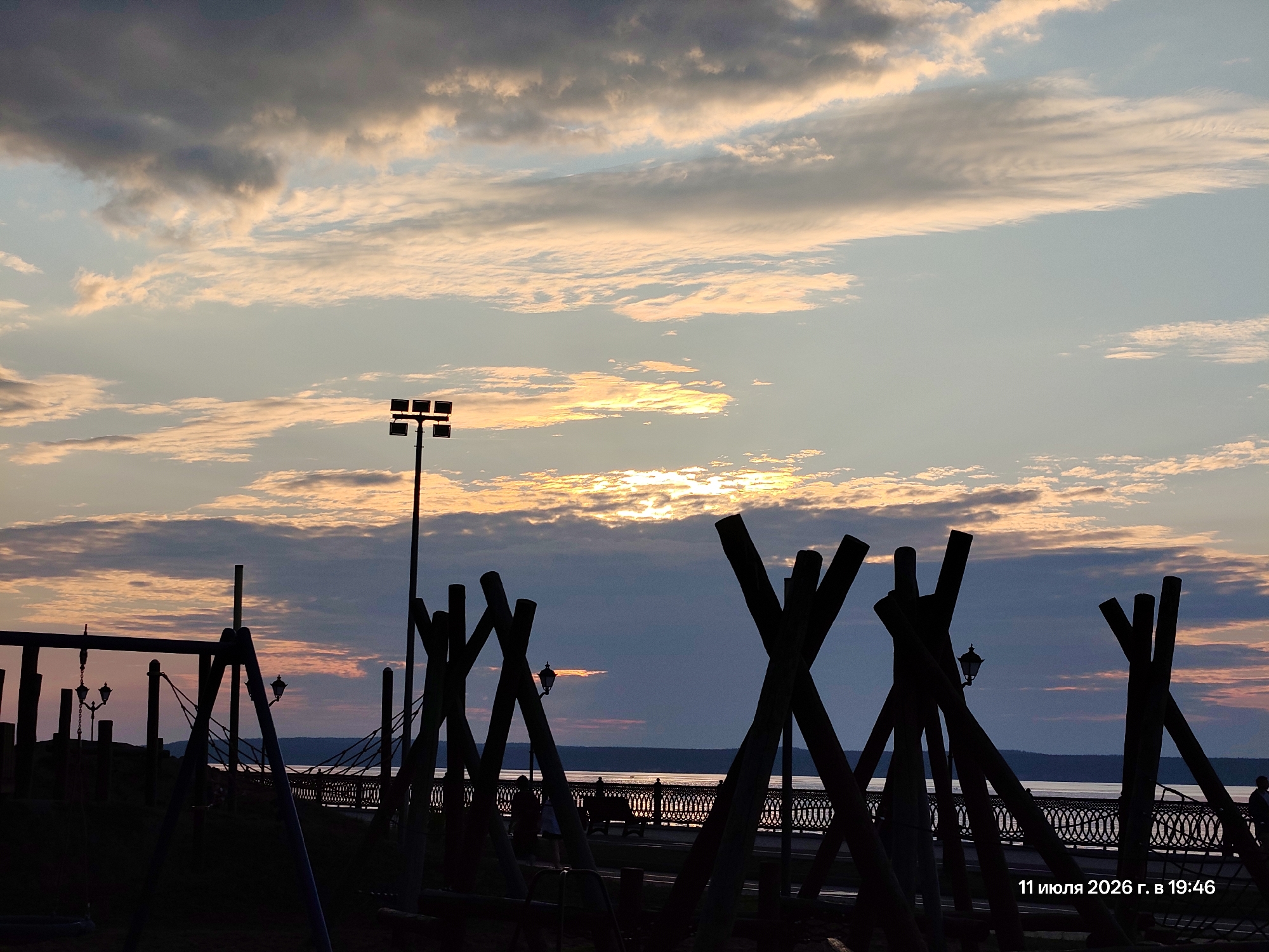 The image captures a waterfront scene at sunset, likely in an urban park or recreational area. Silhouetted against the colorful sky are various structures, including what appears to be a playground with swings and wooden climbing structures resembling teepees or abstract sculptures. A lamppost is visible on the left. In the background, a body of water, possibly a river, stretches across the horizon. Along the water's edge, there's a decorative railing and a walkway where a few indistinct figures are present. The sky is filled with dramatic clouds, with the setting sun casting warm orange and yellow hues, contrasting with darker gray clouds above. The date "11 июля 2026 г. в 19:46" (July 11, 2026, at 19:46) is superimposed in the bottom right corner.