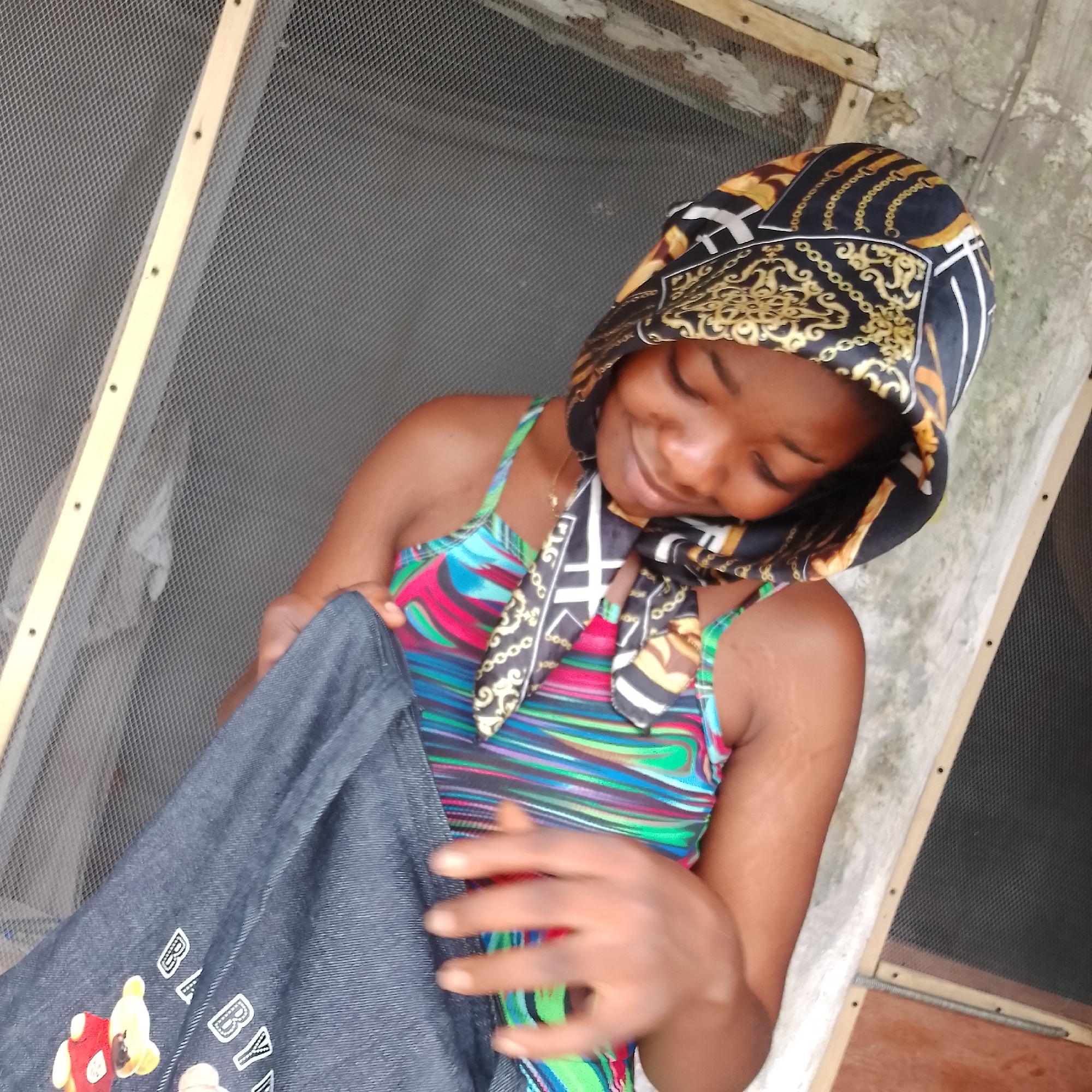 A young Nigerian woman with dark skin and a black headscarf with gold chain and ornate patterns is looking down and smiling. She is wearing a colorful striped dress and holding a dark blue denim jacket. The jacket has embroidered teddy bears and the letters "B" and "Y" visible on it. She is standing in front of a window screen with a wooden frame, and a concrete wall. The scene appears to be outdoors or in a sheltered outdoor area, possibly in a residential setting in Ado, Nigeria.