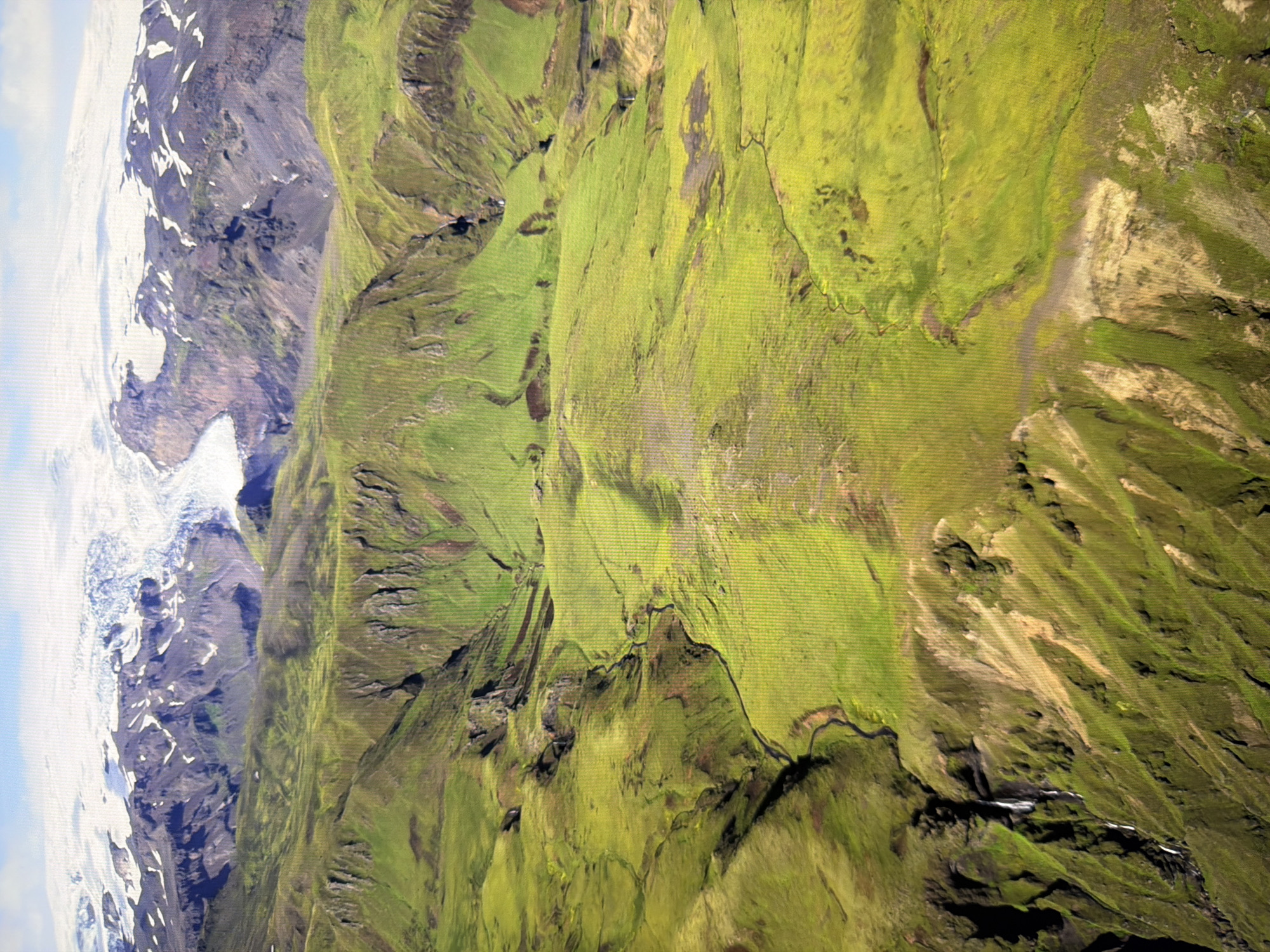 This is an aerial photograph of a vast, mountainous, and glaciated landscape. The foreground is dominated by rolling green hills covered in vibrant moss or grass, with some areas of exposed earth and rock, particularly on steeper slopes. A winding stream or river is visible snaking through the valley. In the middle ground, the terrain becomes more rugged with darker rocky outcrops and steep cliffs. In the background, a large glacier covers the tops of the mountains, appearing white and blue. The sky is a pale blue with some wispy clouds. The image does not depict the city of Kyiv, Ukraine, or any urban environment.