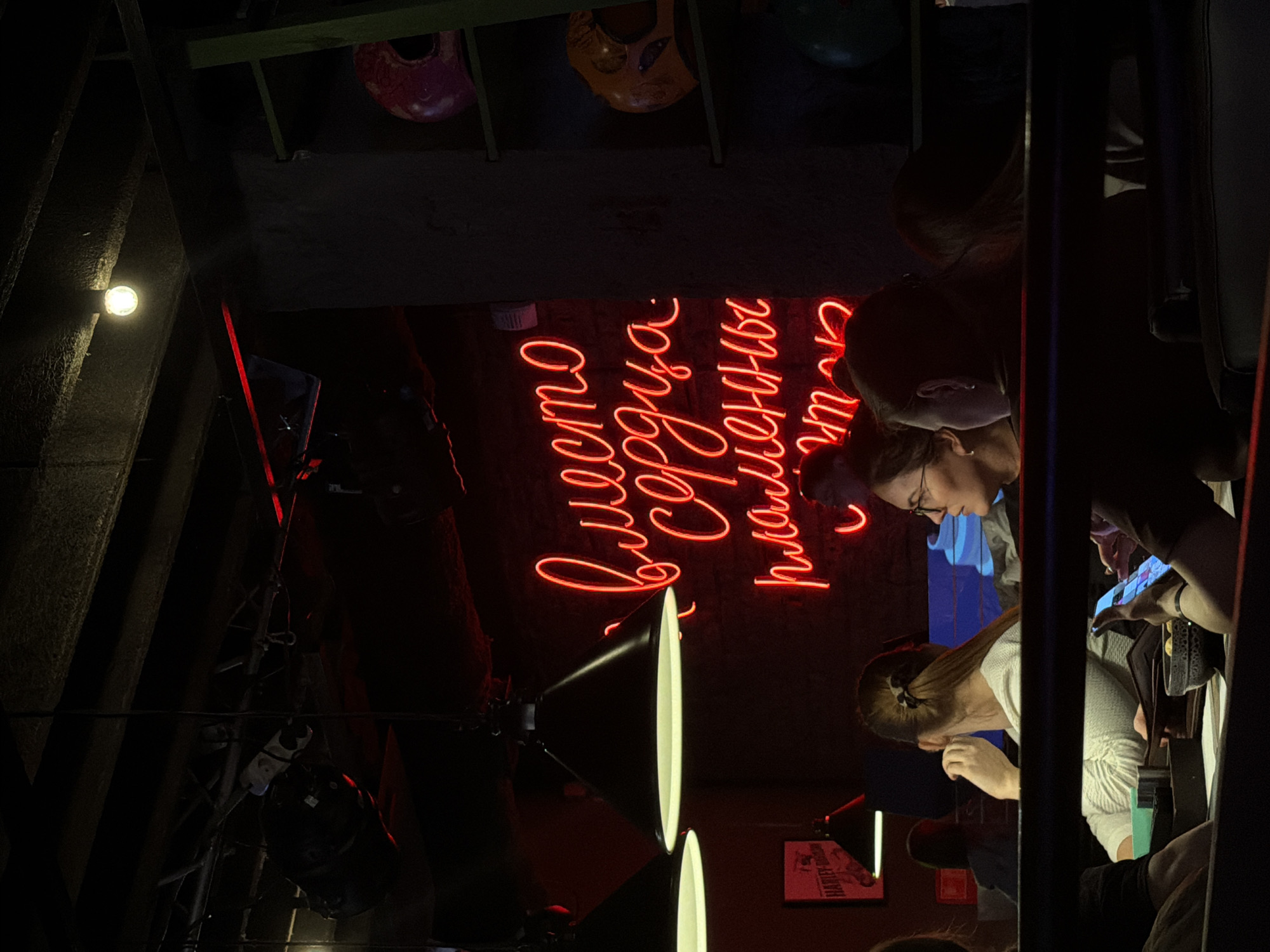 The image depicts the interior of the Harley Davidson bar in Izhevsk, Russia, viewed from a low angle looking up. Two women are visible, seated and engaged with phones or devices, suggesting an interaction or activity involving digital communication. The bar is dimly lit, with neon lighting in the background displaying Russian text. A hanging structure and lighting fixtures are visible in the dark upper portion of the frame.