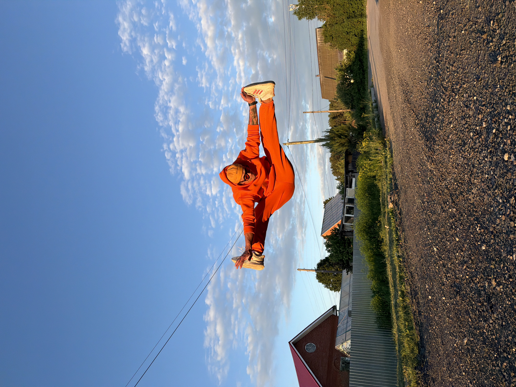 The image, captured in Chaikovsky, Russia, features a person dressed in a vibrant orange hooded sweatshirt and matching sweatpants. The individual, likely male, is captured in a dynamic, upright pose, holding one of their light-colored athletic shoes (which have red stripes) aloft with both hands. Tattoos are visible on their forearms, and a watch is present on one wrist. A cap is worn on their head, obscuring their face.
The scene is set outdoors under a clear blue sky dotted with scattered white cumulus clouds. The background to the right reveals a gravel road, illuminated by the warm, golden light of what appears to be late afternoon or early evening, casting soft shadows. Green trees, bushes, and grass line the road and extend into the distance. Several buildings are visible, including a house with a prominent red roof and another structure with a lighter roof, possibly featuring solar panels. Utility poles and power lines traverse the sky, suggesting a semi-rural environment. No discernible text is visible in the image.