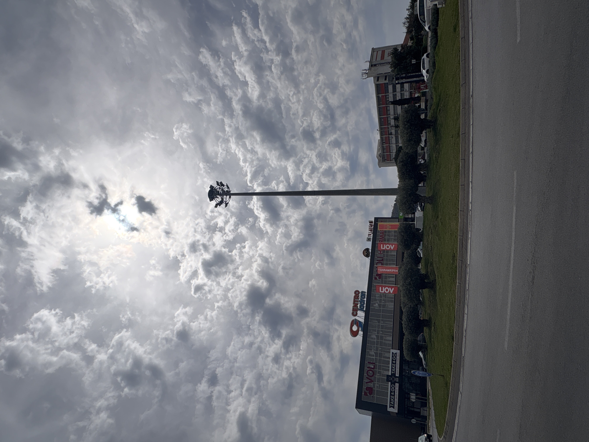 The image is a street-level view, taken horizontally, of a scene in Bar, Montenegro. The lower left portion of the frame features a tall building with "VOLI" signage. To the right, a grassy median divides the building from an asphalt road. The road takes up a significant portion of the frame. A light pole stands prominently between the building and the sky. The sky is filled with altocumulus clouds, with the sun partially obscured. Beyond the building, other buildings can be seen. The scene is overall somewhat overcast.