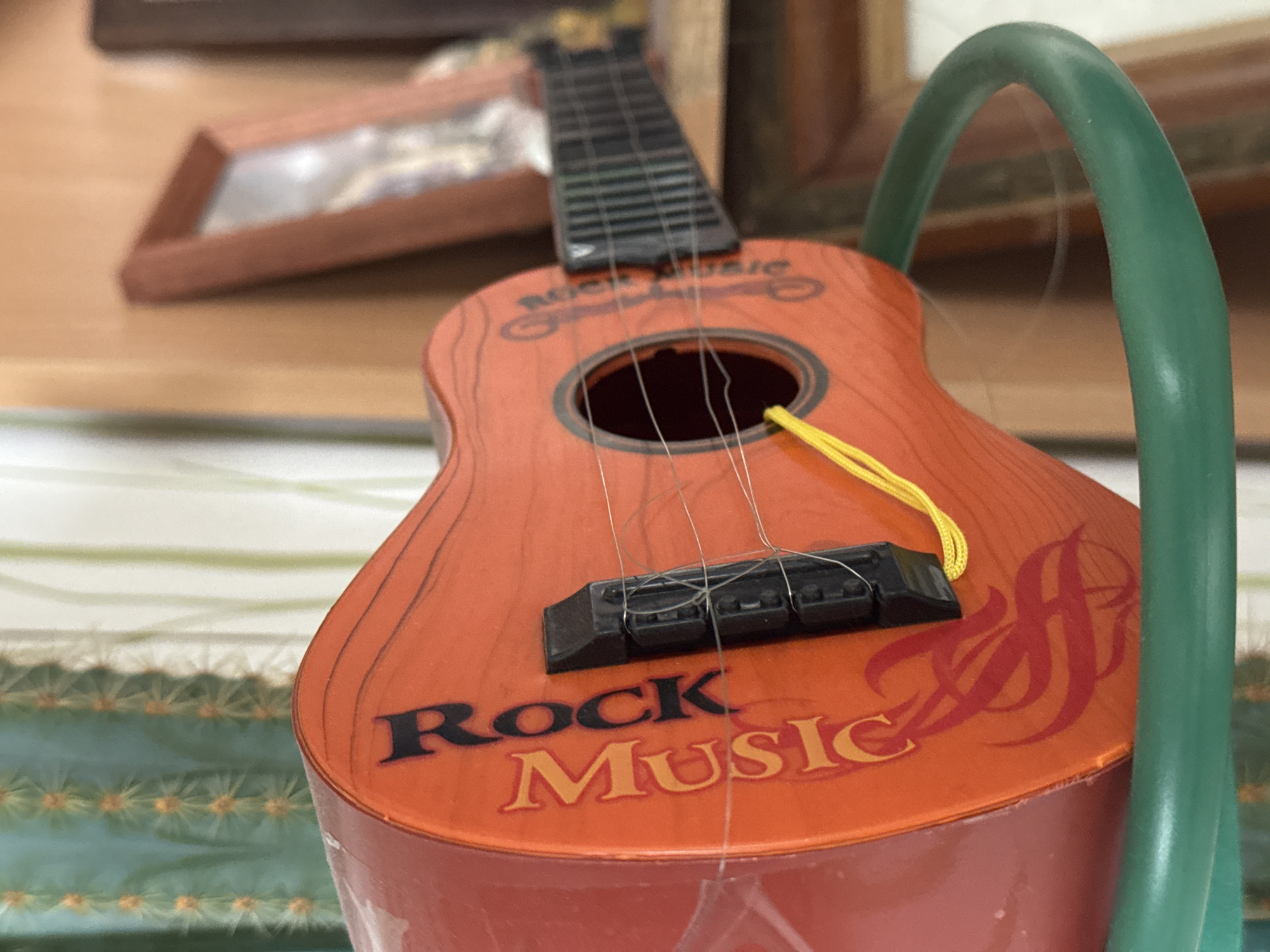 The image features a close-up, shallow depth of field shot of an orange toy guitar with "Rock Music" written on its body. The word "ROCK" is in black capital letters, and "Music" is in a gold-orange color. A stylized red graphic is visible on the right side of the guitar's body. The guitar has a black fretboard and a black bridge, with several thin strings, some appearing loose or broken. A bright yellow string or strap is tied near the bridge and extends towards the right. A prominent curved green object, possibly part of a stand or another toy, is visible on the right side of the frame, partially obscuring the guitar.
The background is blurred, showing soft brown tones of what appears to be a wooden surface, possibly a table or shelf, with a framed object also out of focus. In the lower left, behind the guitar, a blurred green object with numerous small, light-colored spikes is visible, resembling a cactus or similar spiky plant. The scene is indoors, and no specific activity is taking place; the guitar appears to be stationary. The overall impression is a casual, indoor setting, possibly in Novo-Nikolskoye, Russia, as indicated by the provided context.