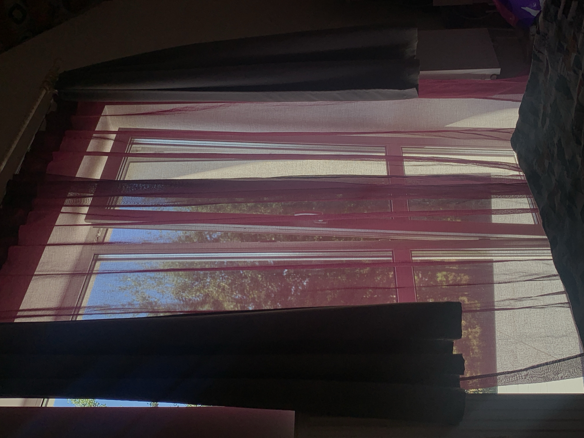 A bedroom is pictured with a large window covered by sheer pink curtains and two opaque dark grey curtains. The window is divided into three vertical panes and is framed in pink. Sunlight streams through the window, illuminating the sheer curtains and casting shadows. Outside the window, trees and a clear blue sky are visible. A bed with patterned bedding is in the foreground, partially obscuring the bottom of the window. The scene is dimly lit, with the exception of the area around the window. The image appears to have been taken indoors, likely in an apartment or house.