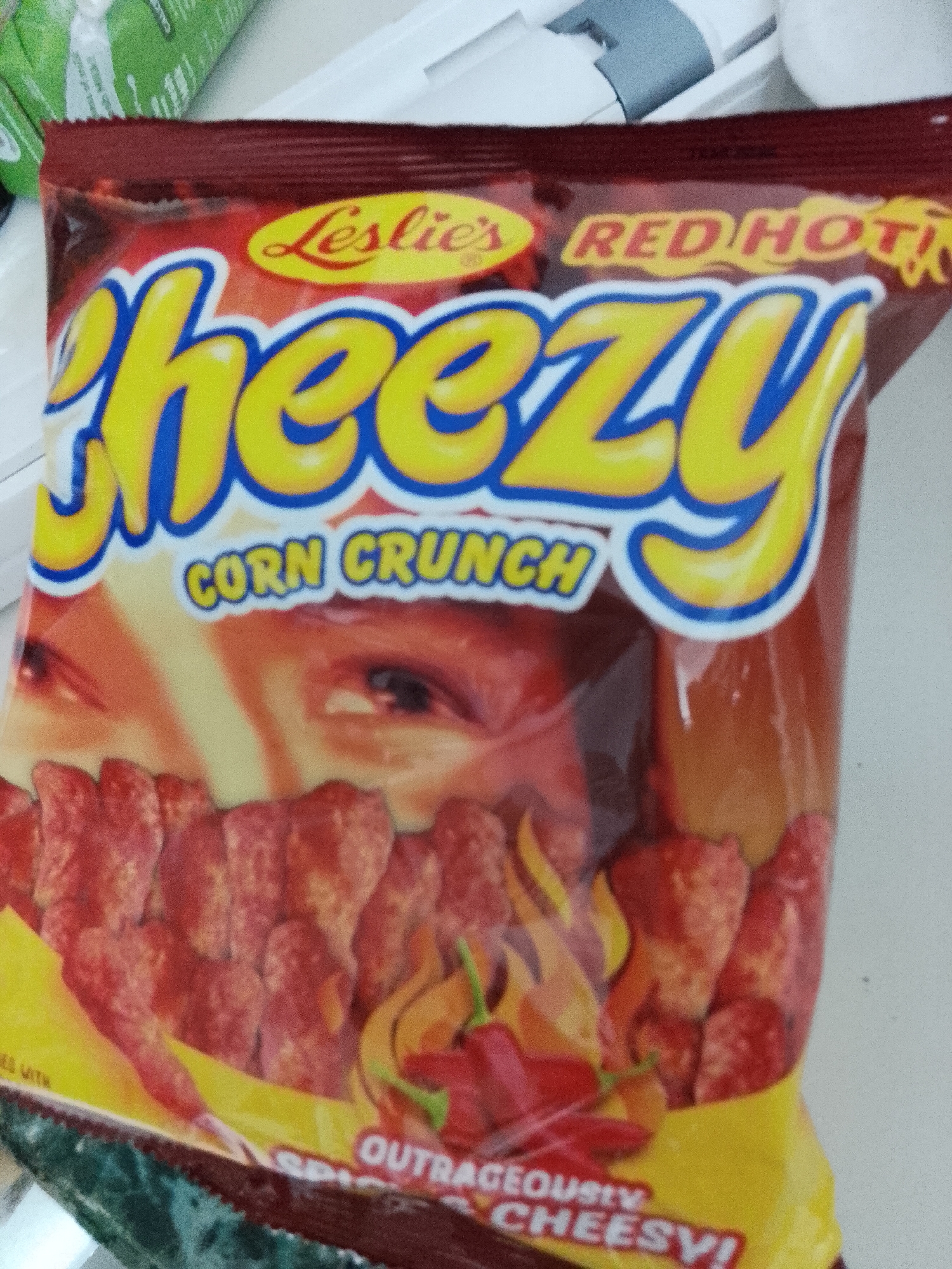 The image displays a vibrant red and yellow snack bag of "Leslie's Cheezy Corn Crunch Red Hot" positioned on a light-colored, possibly white, surface. A portion of green packaging is visible in the upper left background, and another white object or surface is seen behind the snack bag on the right.
The snack bag features prominent text: "Leslie's" in a yellow script font at the top left, "RED HOT!" in yellow with a red outline at the top right, and the large, central text "Cheezy CORN CRUNCH" in bold yellow bubble letters with a blue outline. The packaging design also includes a stylized illustration of a human face, with eyes looking towards the upper right, subtly integrated into the background. Below this, illustrations of numerous elongated, red corn crunch snacks are shown, along with depictions of red chili peppers and orange flames, reinforcing the "Red Hot" claim. Partially visible text at the bottom right of the bag reads "OUTRAGEOUSLY SPICY & CHEESY!".
The scene appears to be an indoor setting, likely a casual environment such as a home, with no specific activity occurring. The presence of a Leslie's brand product is consistent with the given location context of Davao City, Philippines, as it is a well-known Philippine snack brand. The lighting suggests an indoor shot, and no time of day or specific weather is discernible.