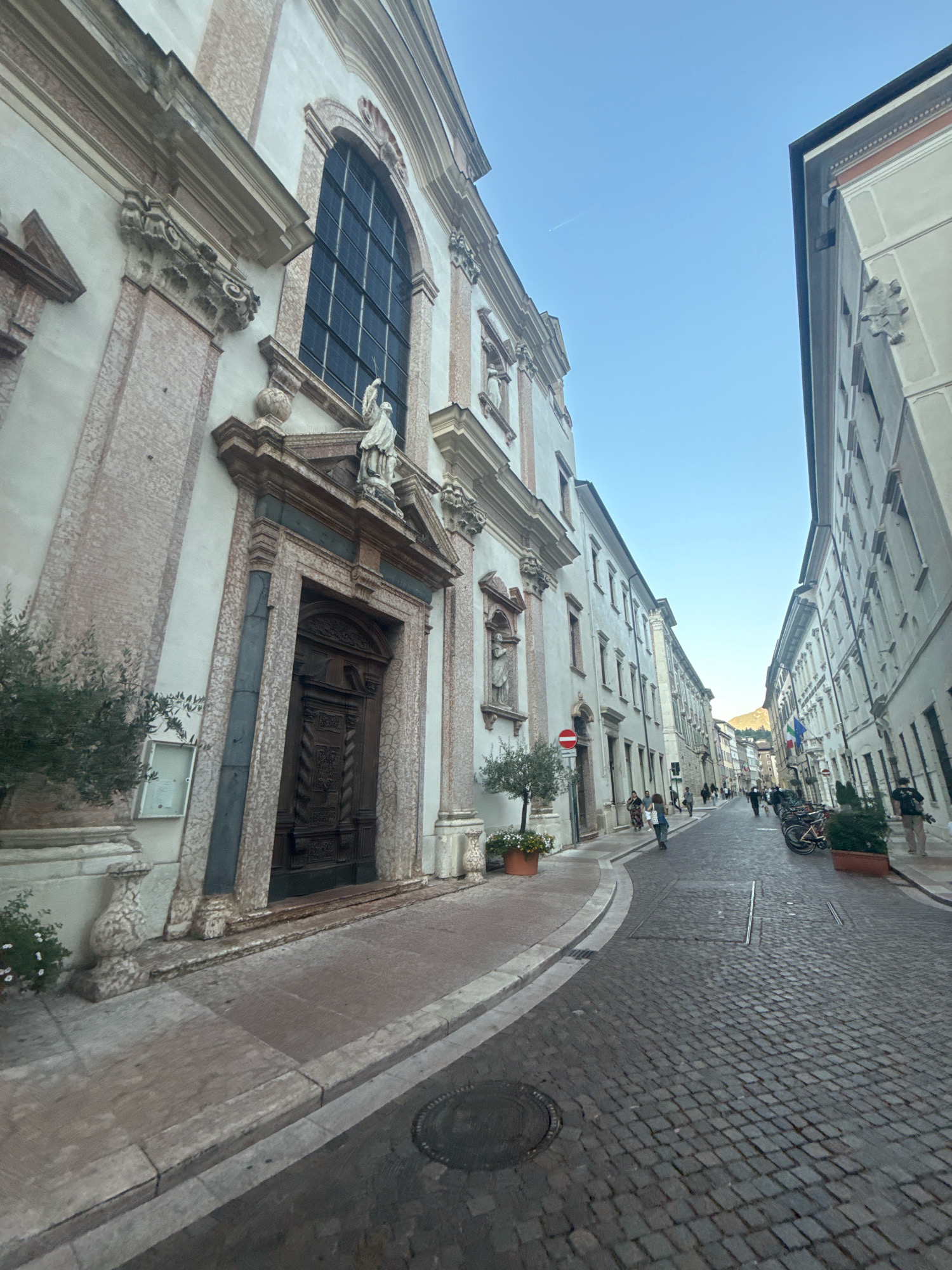 The image shows a street scene in Trento, Italy, with a focus on the facade of a historic building on the left, likely a church or a significant public structure. The architecture is ornate, featuring marble accents, carved wooden doors, and a large arched window. A statue stands above the entrance. To the right of this building, the street curves and is lined with multi-story buildings. The street itself is paved with cobblestones. Several people are visible walking along the street, and bicycles are parked on the right side. The sky is clear and blue, with a contrail from an airplane visible in the upper left quadrant. The scene appears to be during daylight hours, possibly late afternoon given the soft light. The overall impression is of a well-preserved historic urban environment.