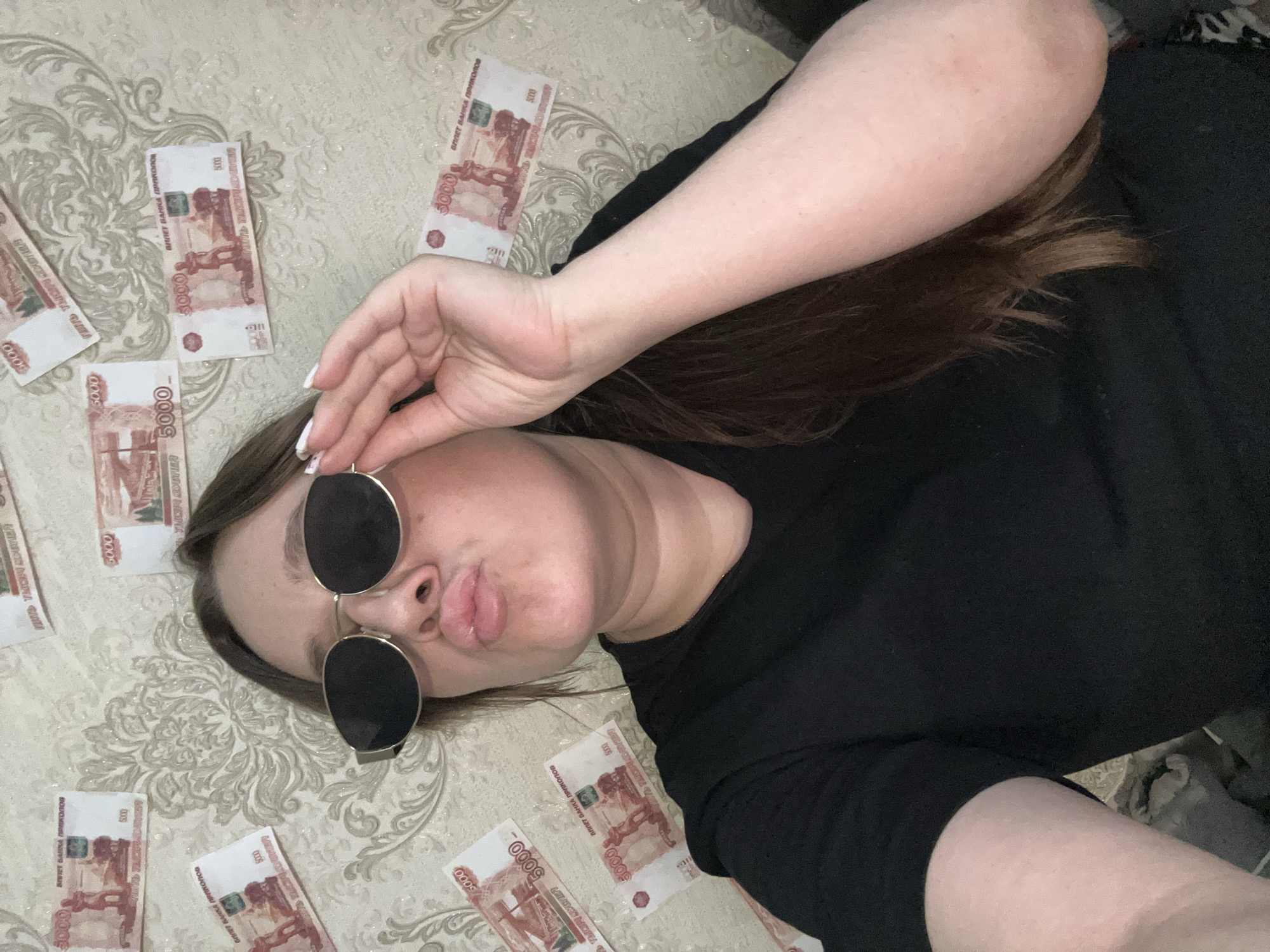 A woman in a black top and sunglasses lies on a patterned surface surrounded by multiple 5000 banknotes. The location is not specified as Chaikovsky, Russia, in the image and cannot be verified.