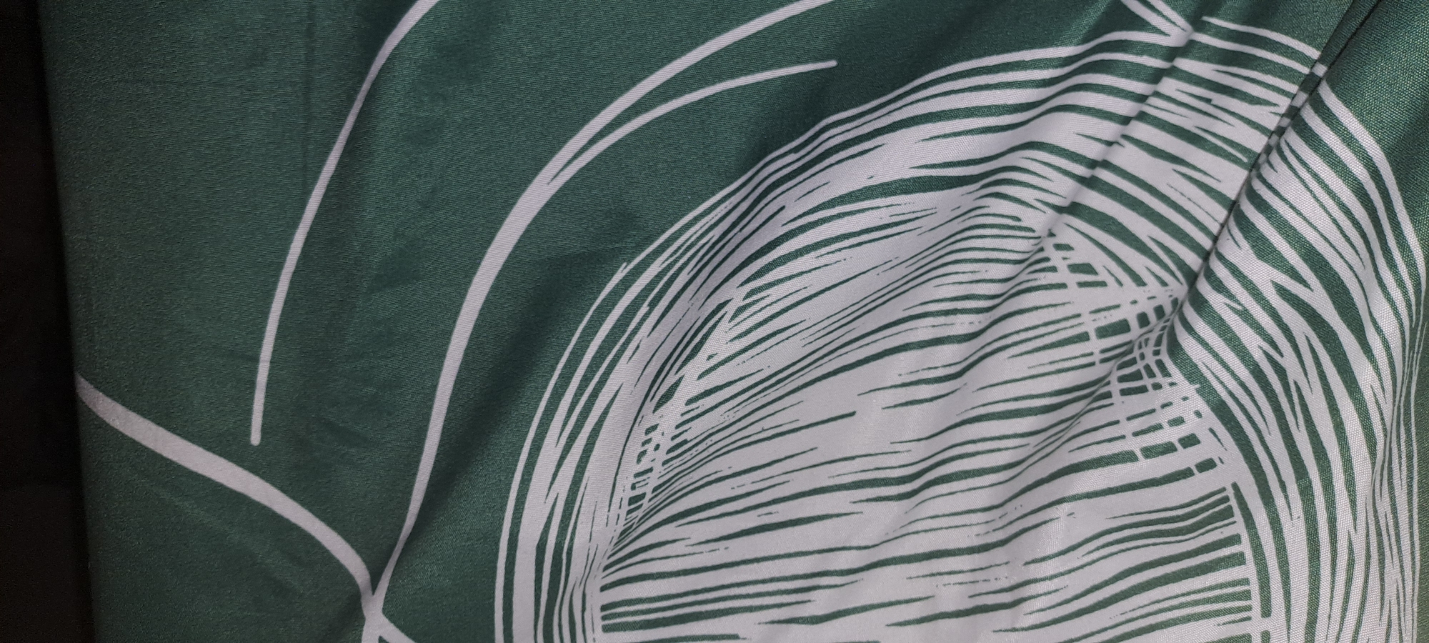 This is a close-up, abstract shot of a green fabric with a white, stylized palm leaf pattern. The fabric is folded and creased, creating shadows and highlights that emphasize the texture and the curves of the design. The palm leaf motif is rendered in bold white lines against the deep green background, with some of the lines appearing thin and delicate, while others are thicker and more solid. The image has a soft focus, adding to the artistic and textured feel. There is no discernible activity, time of day, weather, location cues, or visible text.