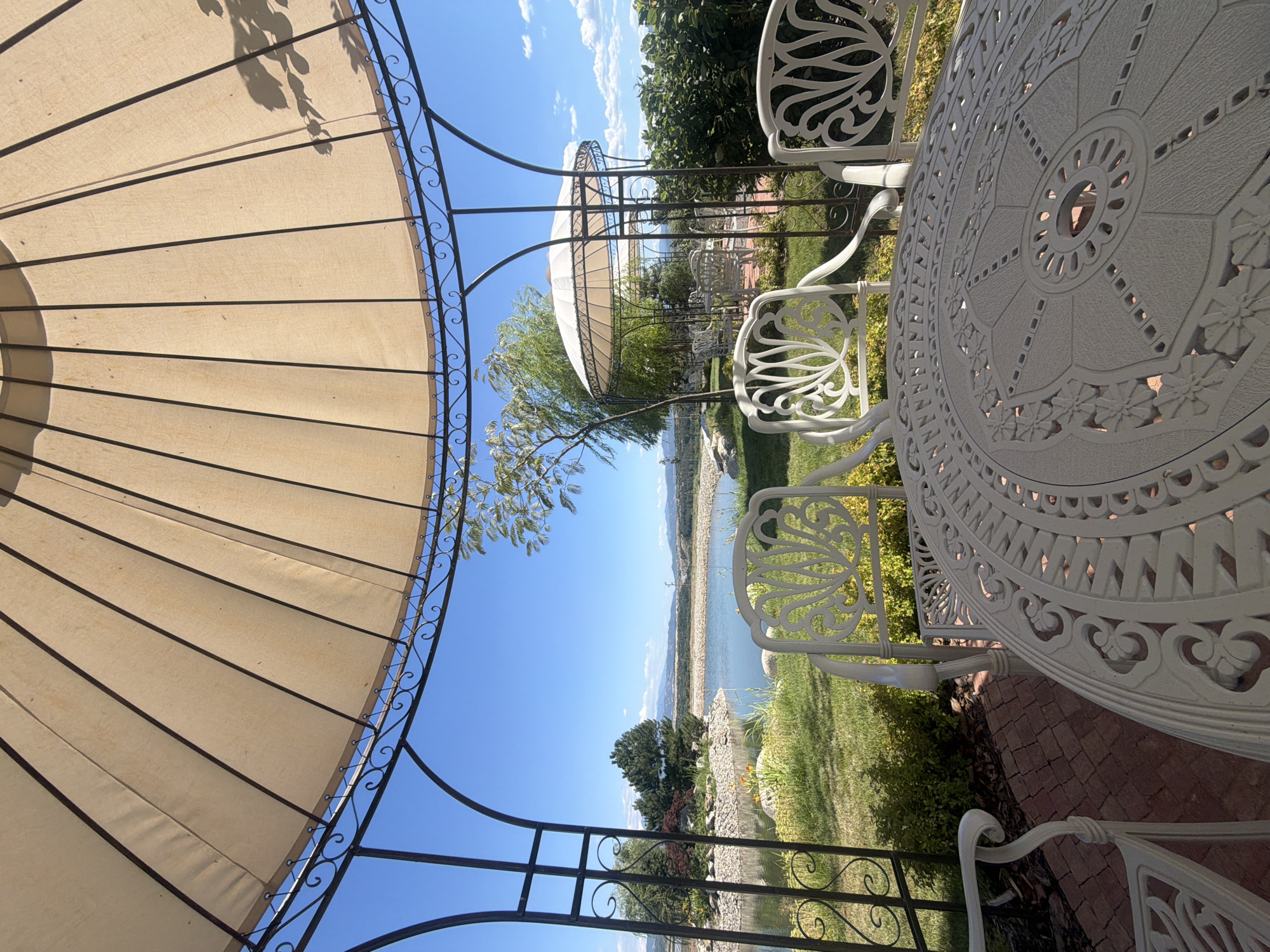 The image depicts a picturesque outdoor seating area beneath a beige gazebo, overlooking a serene lake and rolling hills under a clear blue sky. In the foreground, a white wrought-iron table with an ornate circular design is set, accompanied by matching chairs with intricate scrollwork. The gazebo's canopy casts a shadow, dappling the scene. Beyond the seating area, a well-maintained lawn leads to the edge of the lake, which is surrounded by a rocky embankment and lush greenery. Trees, including a weeping willow, dot the landscape. In the distance, mountains form a hazy horizon. The setting suggests a tranquil environment, possibly a park, resort, or private garden.