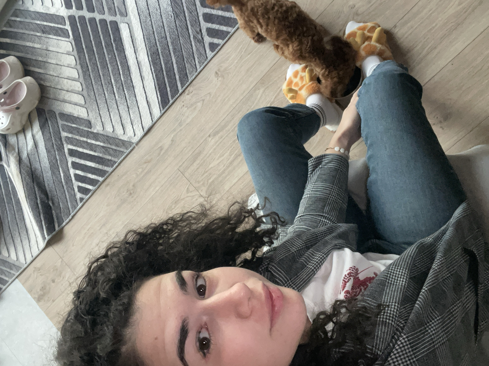 A young woman with curly dark hair is lying on her back on a light-colored wooden floor, wearing blue jeans, a white t-shirt, and a grey plaid blazer. She is looking at the camera. On her feet, she is wearing white socks and orange giraffe-themed slippers. Next to her feet is a small, brown, curly-haired dog. In the background, there is a grey and white patterned rug, and a pair of white shoes. The location appears to be indoors, possibly a home. There is no information to determine the specific location within Sofia, Bulgaria.