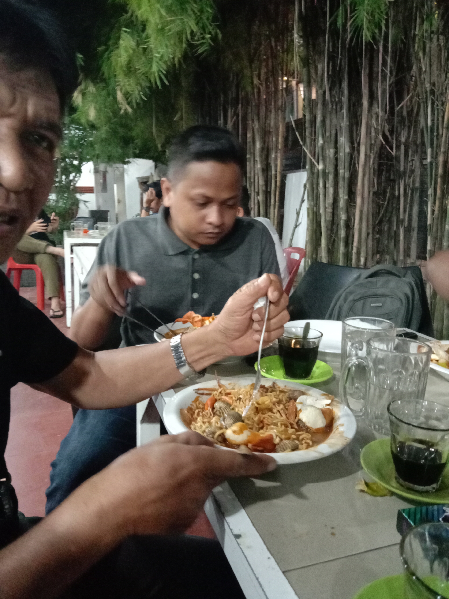 Three men sit at a table outside a restaurant in Lhokseumawe, Indonesia. One man wears a black t-shirt and a silver watch. Another man is seated next to him and is wearing a grey polo shirt. Both men have plates of food in front of them consisting of noodles, shrimp, and what looks like eggs. A third man is seated to the left and appears to be looking at the camera. The table has several glasses of water and a glass of coffee. Behind the men is a decorative bamboo wall and some foliage. Other patrons sit at tables in the background.